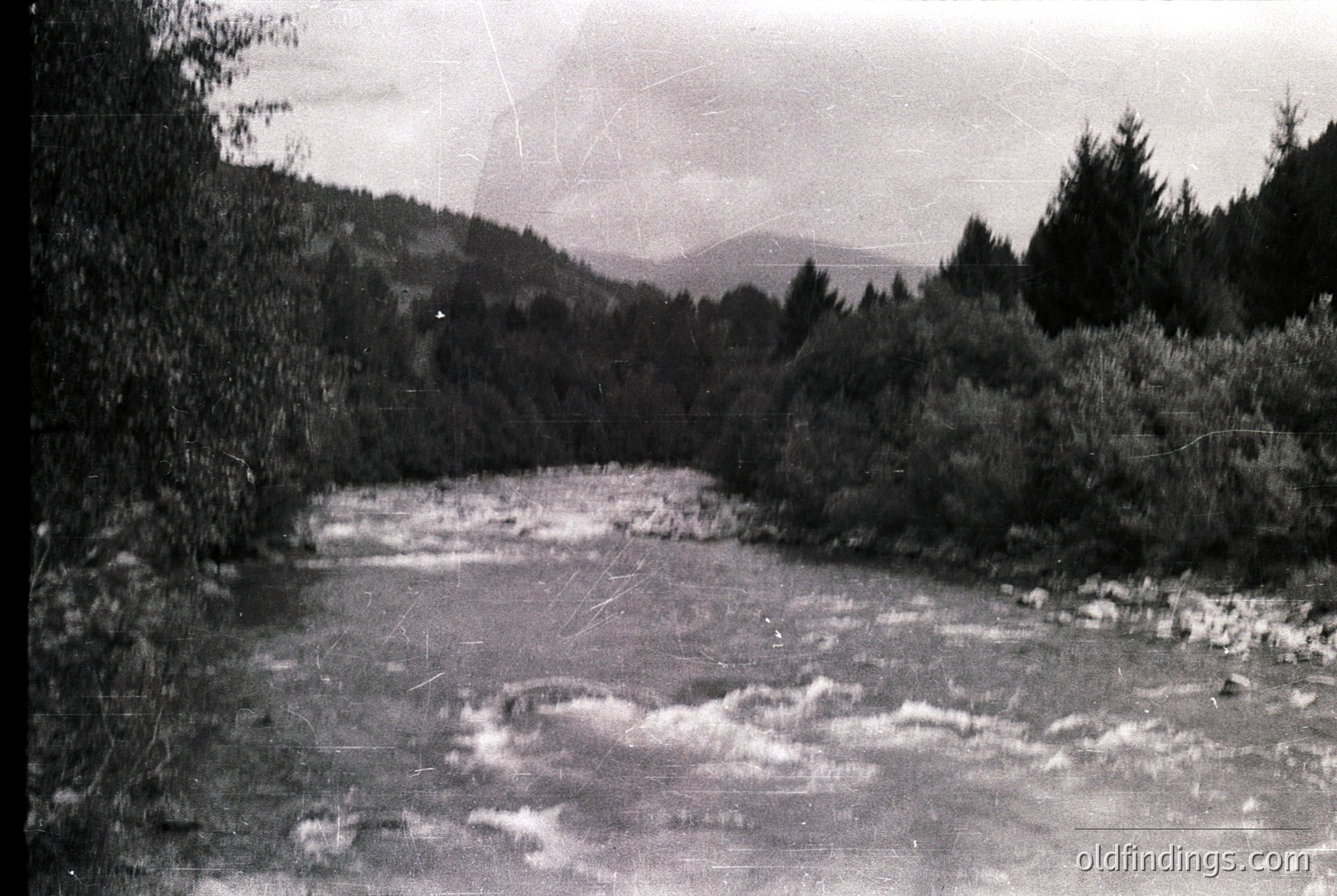 Vintage black-and-white shot of a shallow, rocky river flowing through a forested valley. Dense coniferous trees frame both sides, with mist rising from the water. Distant mountains form a hazy backdrop under an overcast sky. Likely mid-20th century, possibly for nature/landscape documentation or historical research.