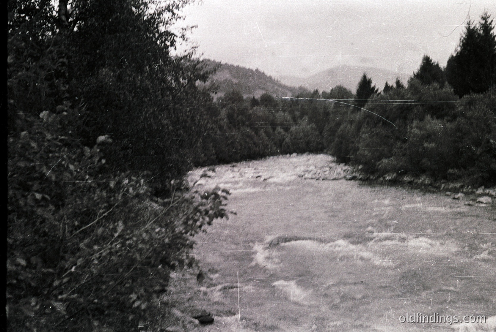 Vintage black-and-white shot of a turbulent river flowing through a forested valley, framed by dense foliage on both sides. Rolling hills and distant mountains create a layered landscape. Likely mid-20th century due to grain and contrast.