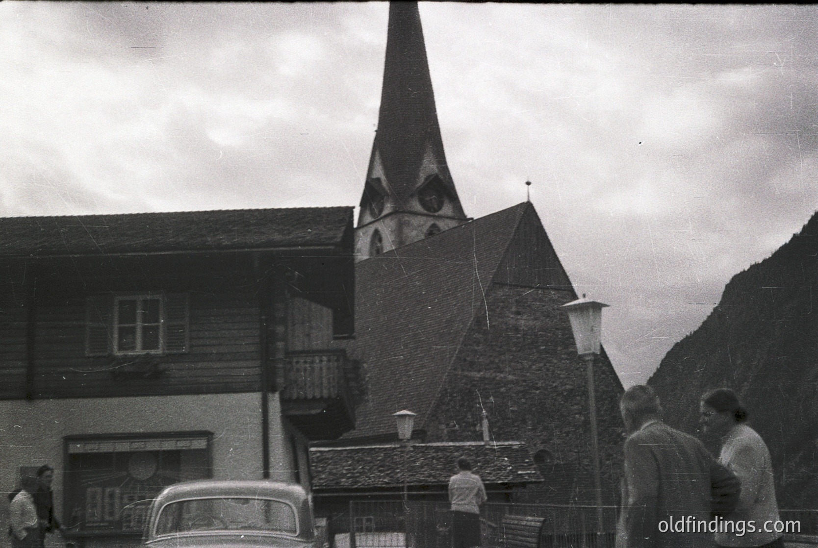Black-and-white alpine village scene featuring a timber-framed chalet and stone church with a steep spire. Classic vintage car parked near a lamppost, with three men standing in conversation. Mountain backdrop suggests mid-20th century European alpine architecture.