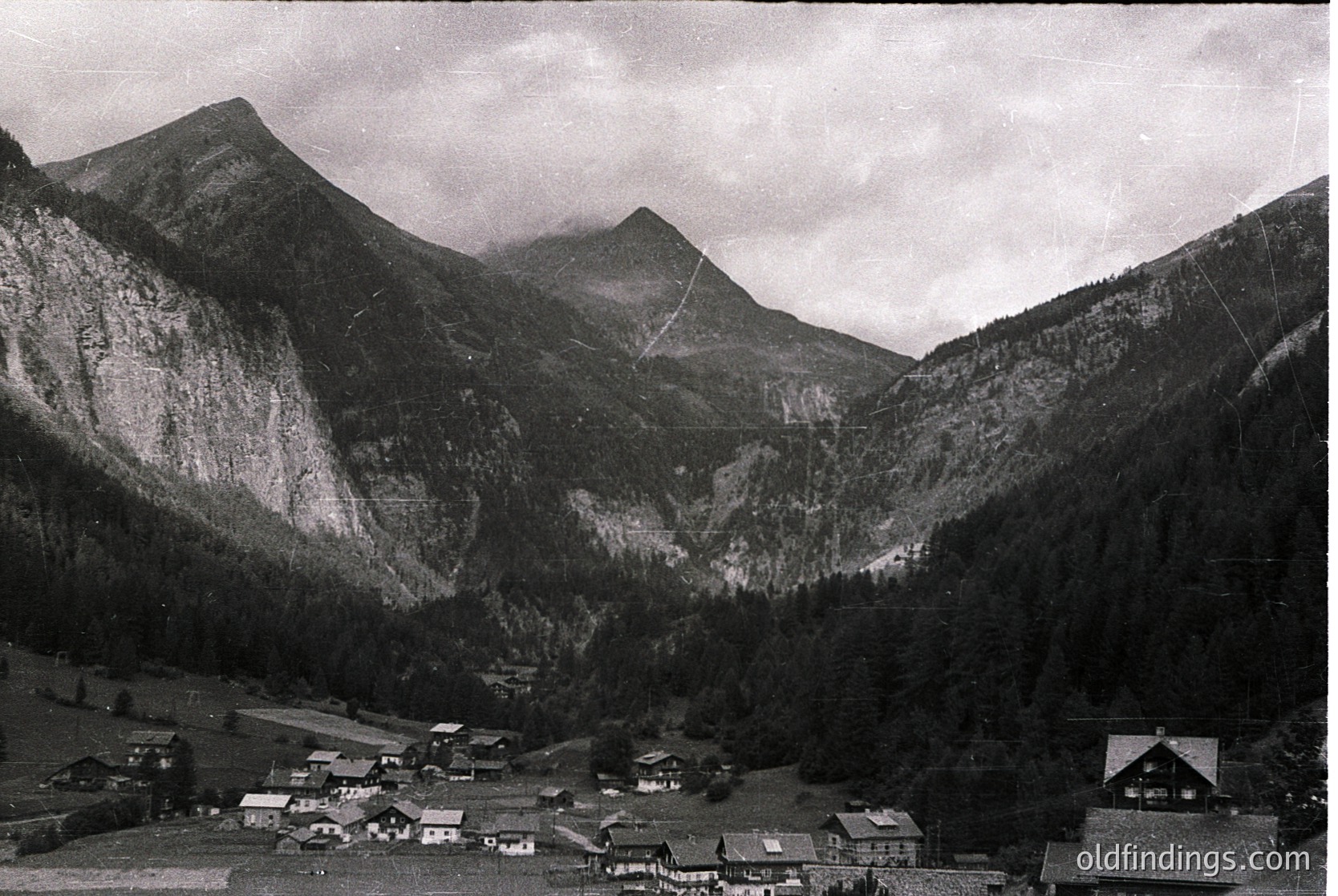 Mid-20th century alpine village nestled between steep, forested slopes and jagged peaks. Classic wooden chalets with pitched roofs cluster along a winding valley road. Dramatic cloud cover enhances the rugged mountain backdrop.