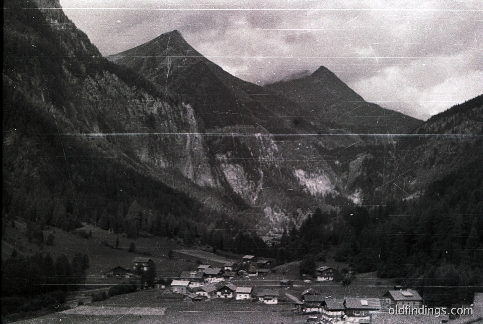 Aerial black-and-white view of a mountainous Alpine village nestled in a valley, likely mid-20th century. Clustered wooden chalets with sloped roofs and narrow streets winding through dense forest. Dramatic peaks and steep slopes dominate the background, suggesting rugged terrain. Ideal for historical research or alpine-themed design references.
