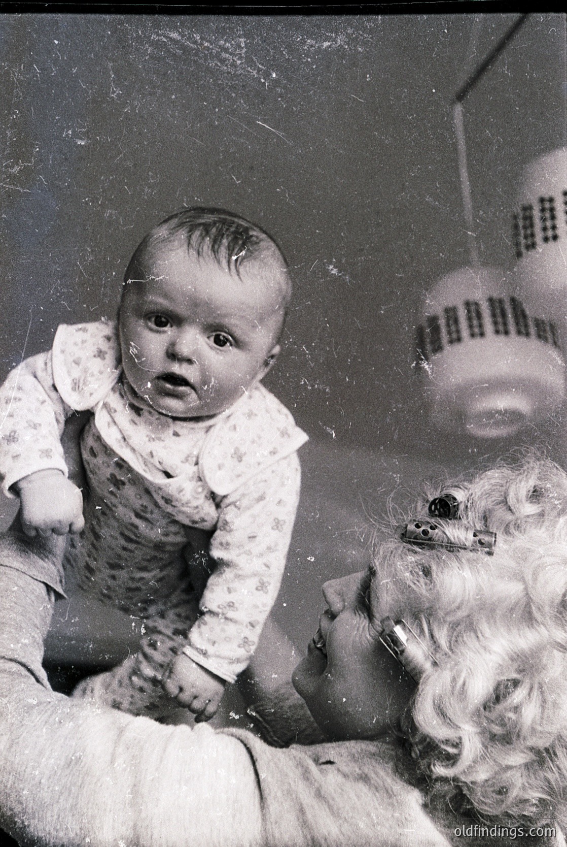 Vintage black-and-white photo of an infant in a bathtub, surrounded by soapy water and bubbles. The child wears a floral-patterned onesie, and a woman’s arm (with a watch) supports them. A vintage hairdryer with a grid vent sits on the edge. Mid-20th century domestic life captured.