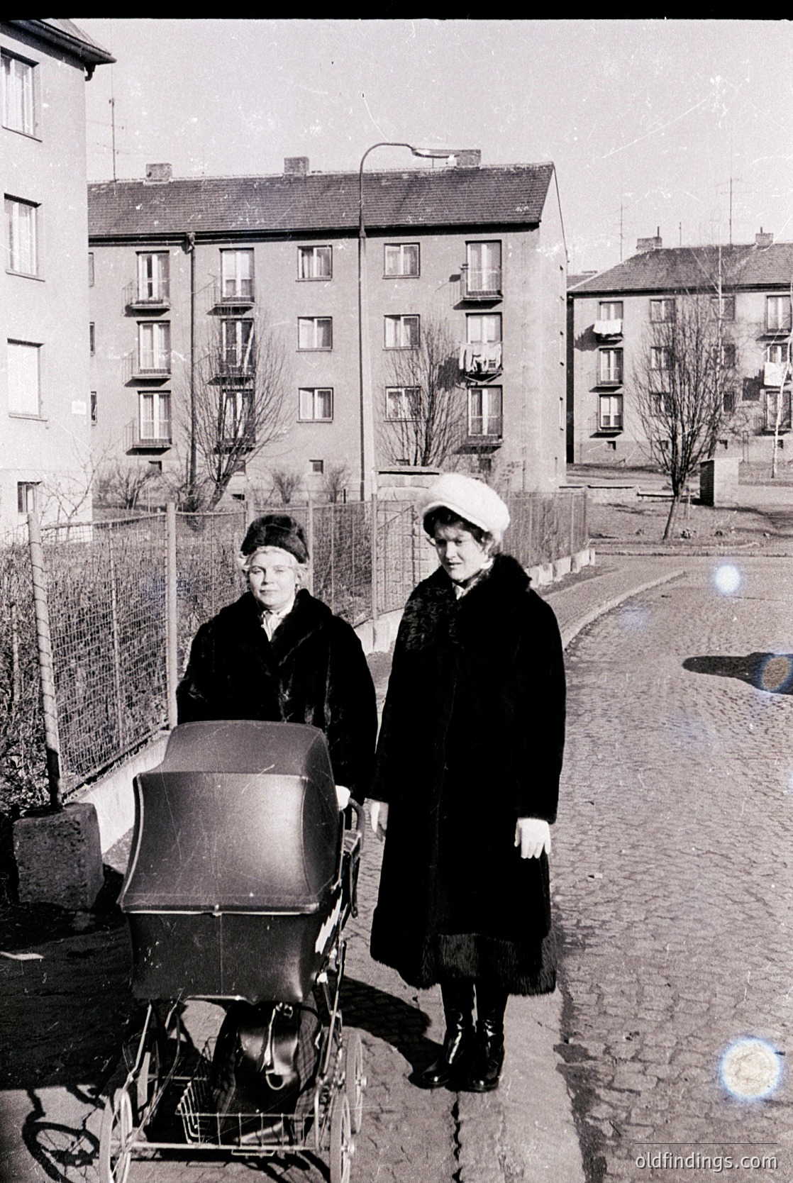Two women in 1950s-60s winter attire stand beside a vintage pram on a cobblestone street. Brick apartment buildings with balconies and leafless trees frame the scene, suggesting urban Eastern Europe. The women’s coats and headscarves reflect mid-century fashion.