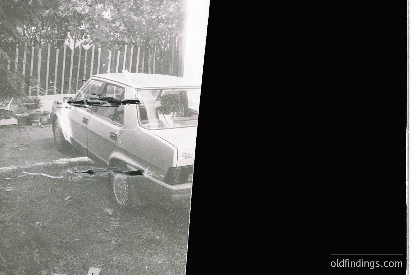Vintage black-and-white photo of a classic sedan parked on uneven terrain, likely post-1960s. Vehicle appears to be a European model, possibly a Lada or similar Soviet-era car, with visible rust and damage. Surrounding area shows overgrown vegetation and a wooden fence, suggesting rural or suburban setting. Rain or snow partially obscures visibility.