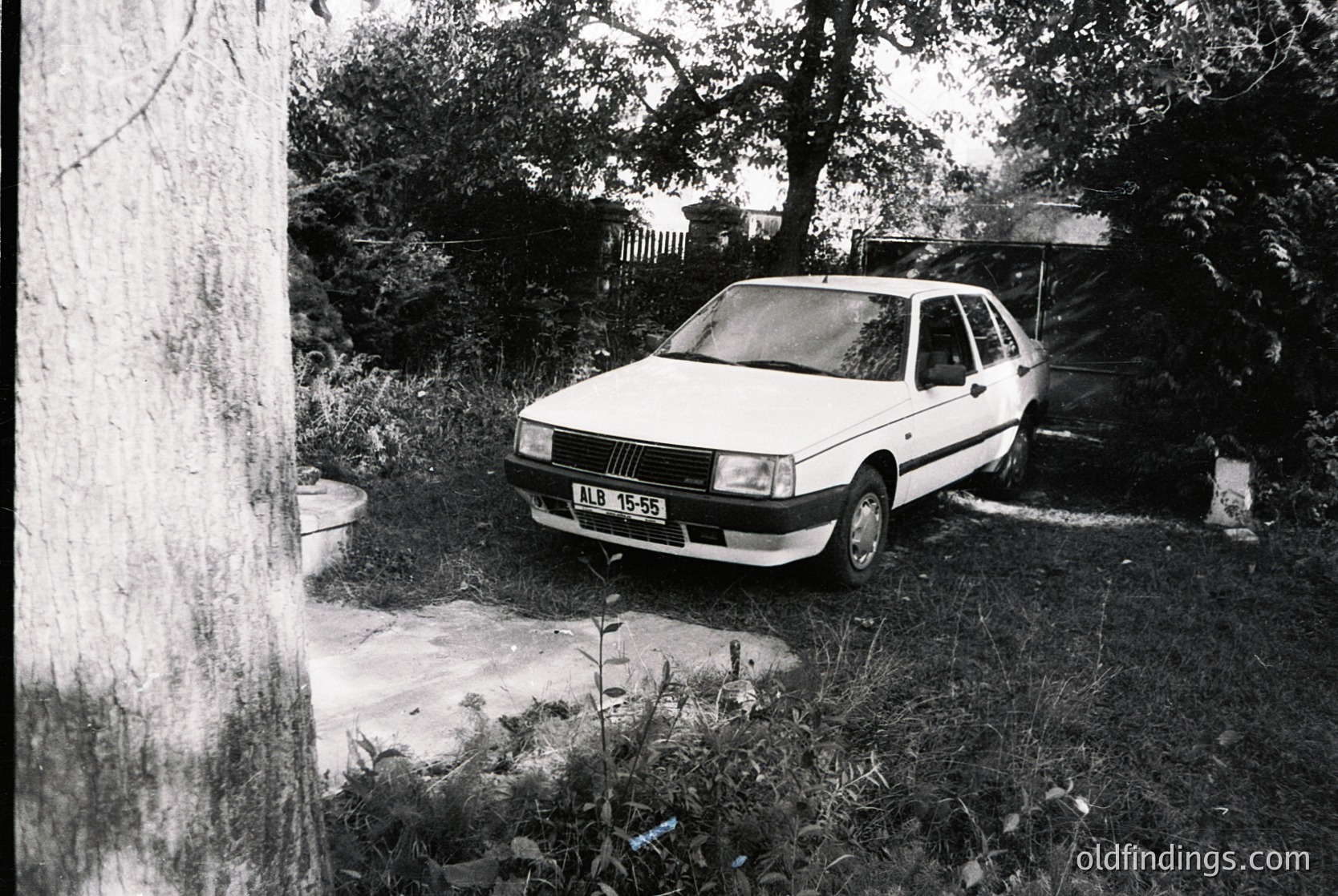 Classic **Fiat 128** (1970s model) parked in a rural or suburban driveway, framed by overgrown foliage and a tree trunk. Black-and-white photo captures mid-century automotive design with minimalist license plate "AB 19-55." Rustic, nostalgic setting suggests European countryside or small town.
