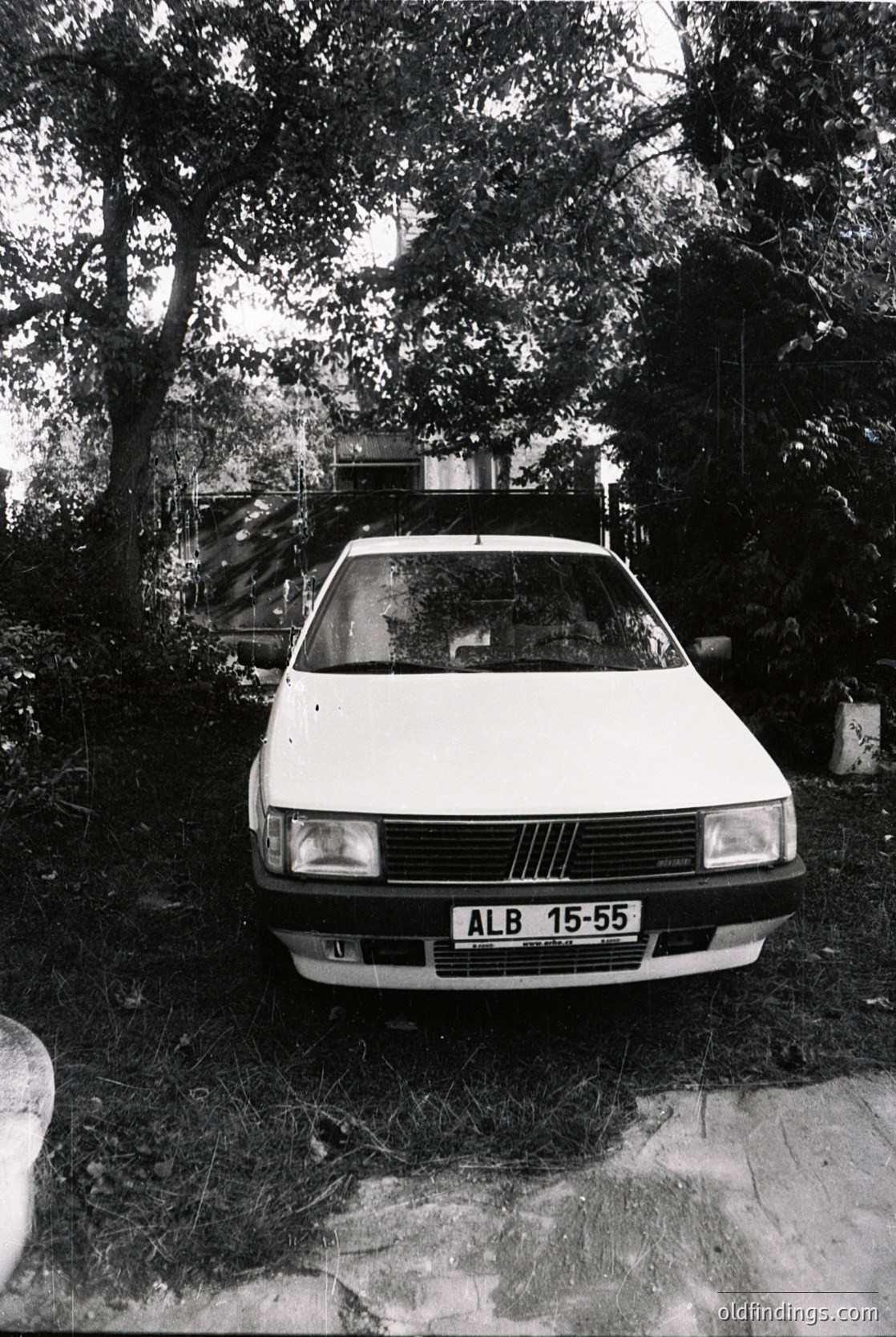 Vintage Fiat 128 sedan (ALB 15-55 license) parked in a wooded driveway, surrounded by dense foliage. Mid-20th century European design with classic rectangular headlights and vertical grille. Overgrown grass and stone pathway suggest rural or suburban setting.