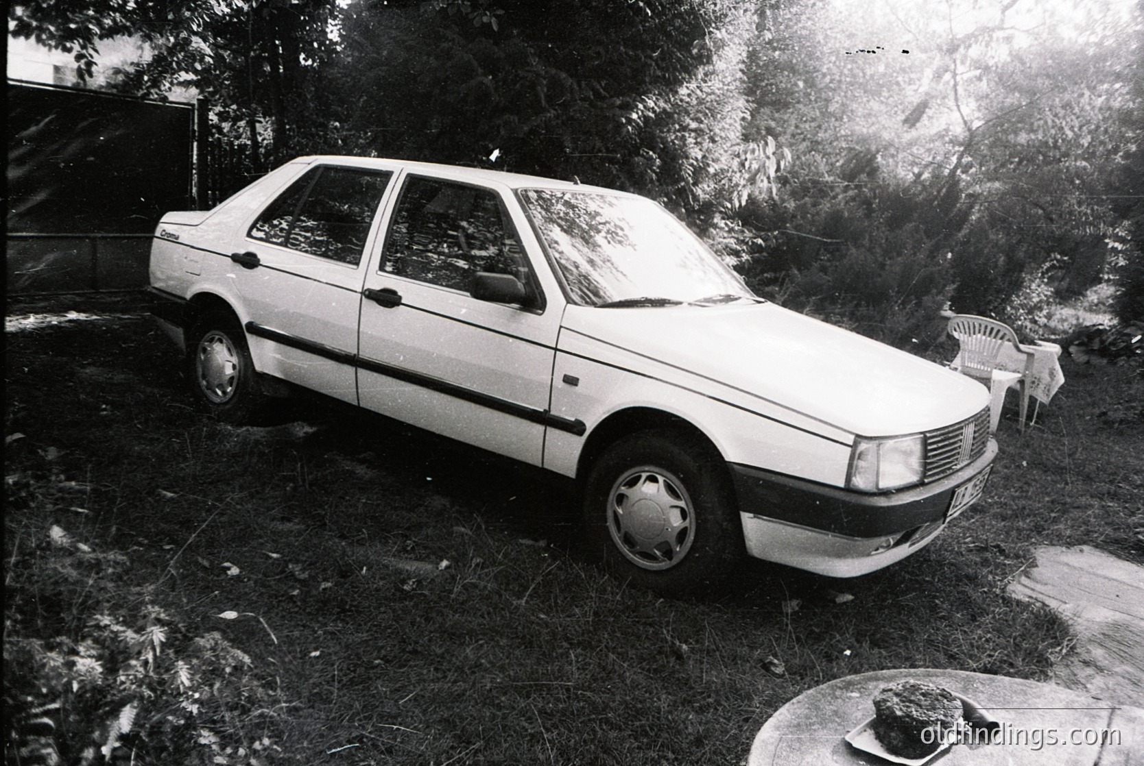 Classic sedan parked on grassy yard, likely a **Volkswagen Passat** (B1/B2 generation, 1980s). Rusting metal, faded paint, and worn tires suggest mid-to-late 1980s European setting. Side mirror and rearview mirror reflect autumn foliage.