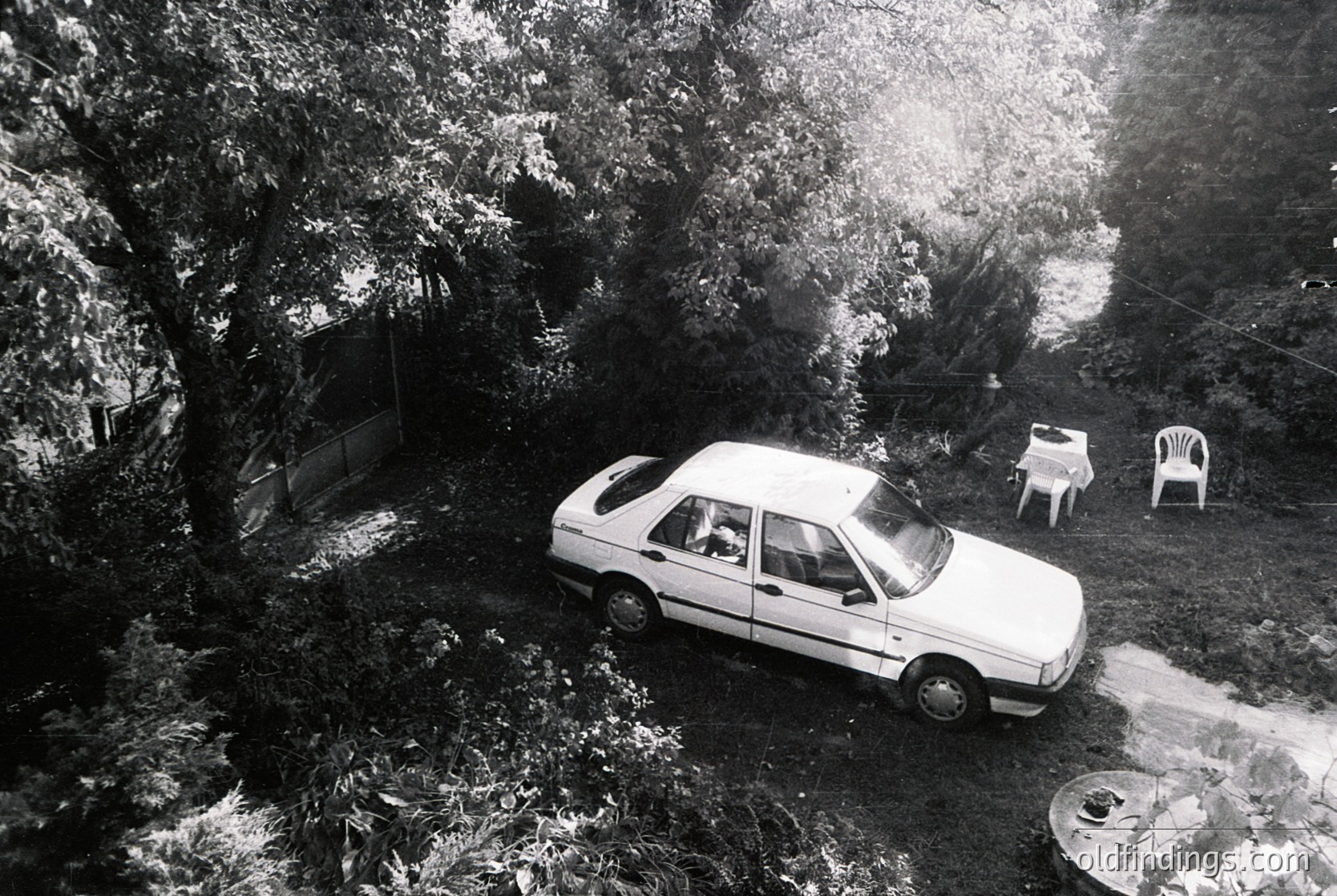 Aerial shot of a 1980s-era sedan parked on a sloped driveway surrounded by dense foliage. Two white plastic chairs and a table set on grass beside a small wooden shed. Snow blankets the ground, indicating a winter scene.