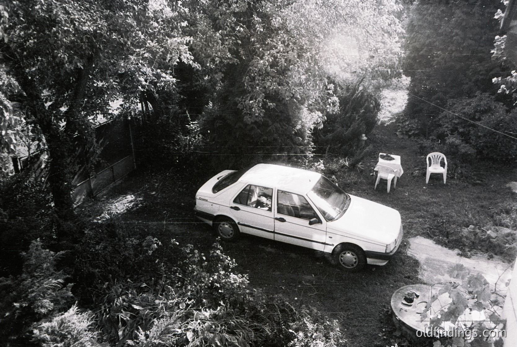 Aerial view of a mid-20th-century sedan parked in a shaded backyard, surrounded by dense foliage. Two white plastic chairs and a table sit near a wooden structure, likely a shed or small building. The scene evokes a midcentury suburban or rural setting, possibly or .