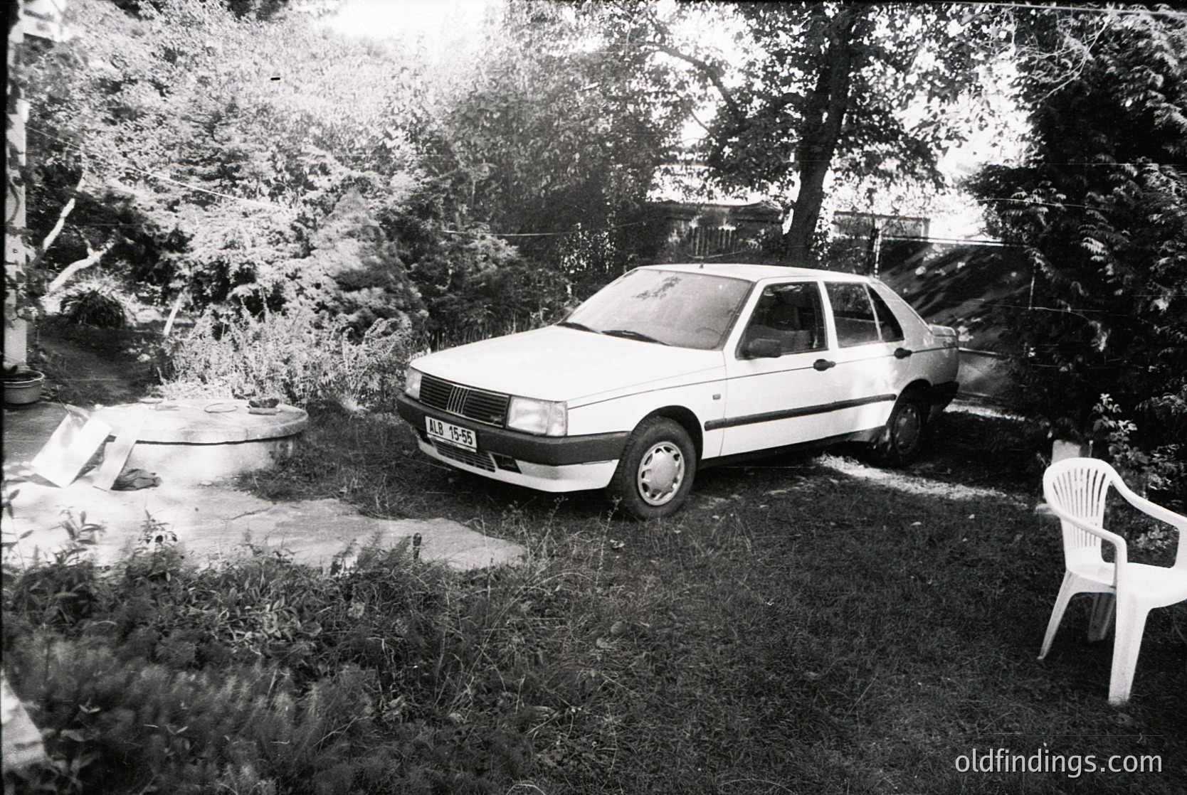 Vintage sedan parked on overgrown grass beside a concrete slab and plastic chair. Likely a 1980s European model, suggested by design and license plate format. Residential yard with mature trees and rustic surroundings.