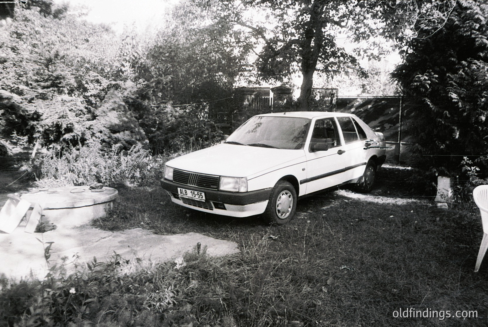 A 1980s-era Fiat sedan parked in a residential backyard, surrounded by overgrown foliage. Concrete steps and a concrete planter frame the scene, suggesting mid-century suburban design. Black-and-white monochrome enhances vintage aesthetic.