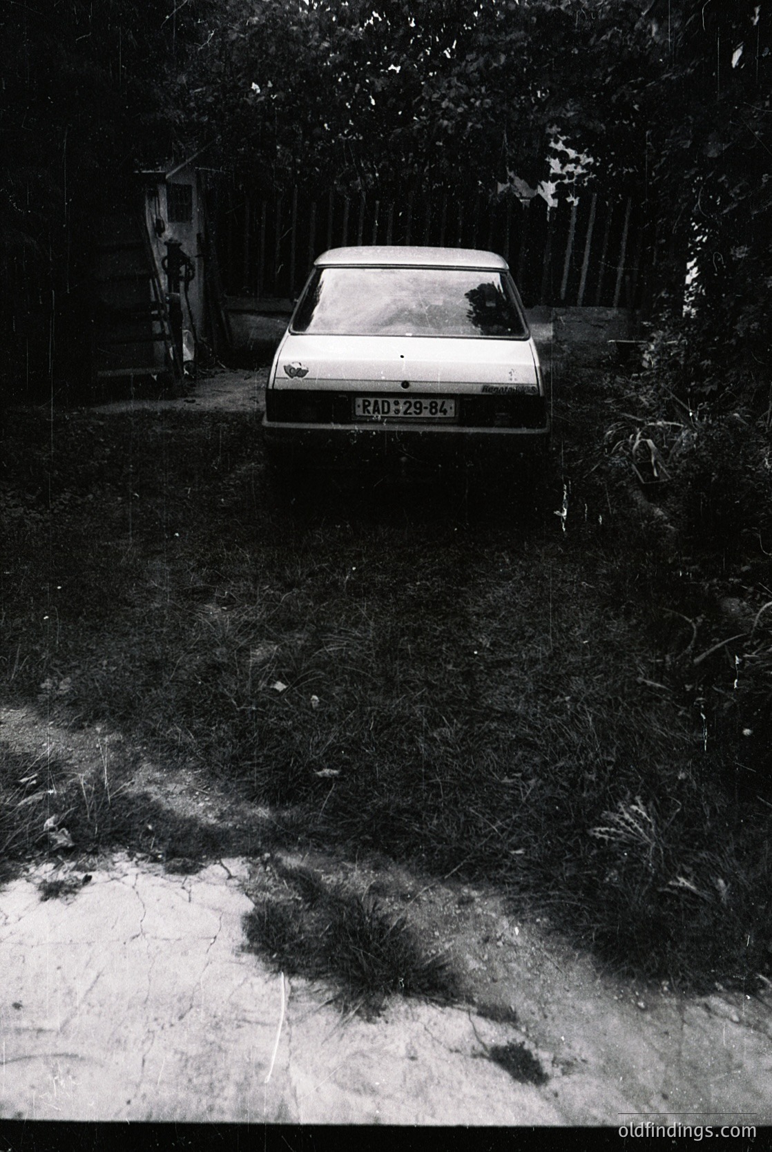 Vintage sedan parked on unpaved driveway beside overgrown foliage; license plate "RD 25-84" suggests Eastern Bloc-era vehicle, likely 1970s–1980s. Rustic, rural setting with wooden fence and shadowed pathway.