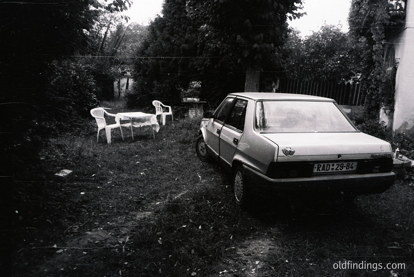 Vintage black-and-white shot of a neglected suburban yard: a faded white sedan (RAD-29-84) parked beside overgrown grass, with a plastic chair and table set abandoned nearby. Residential fence and trees frame the scene, suggesting mid-20th century domestic life.