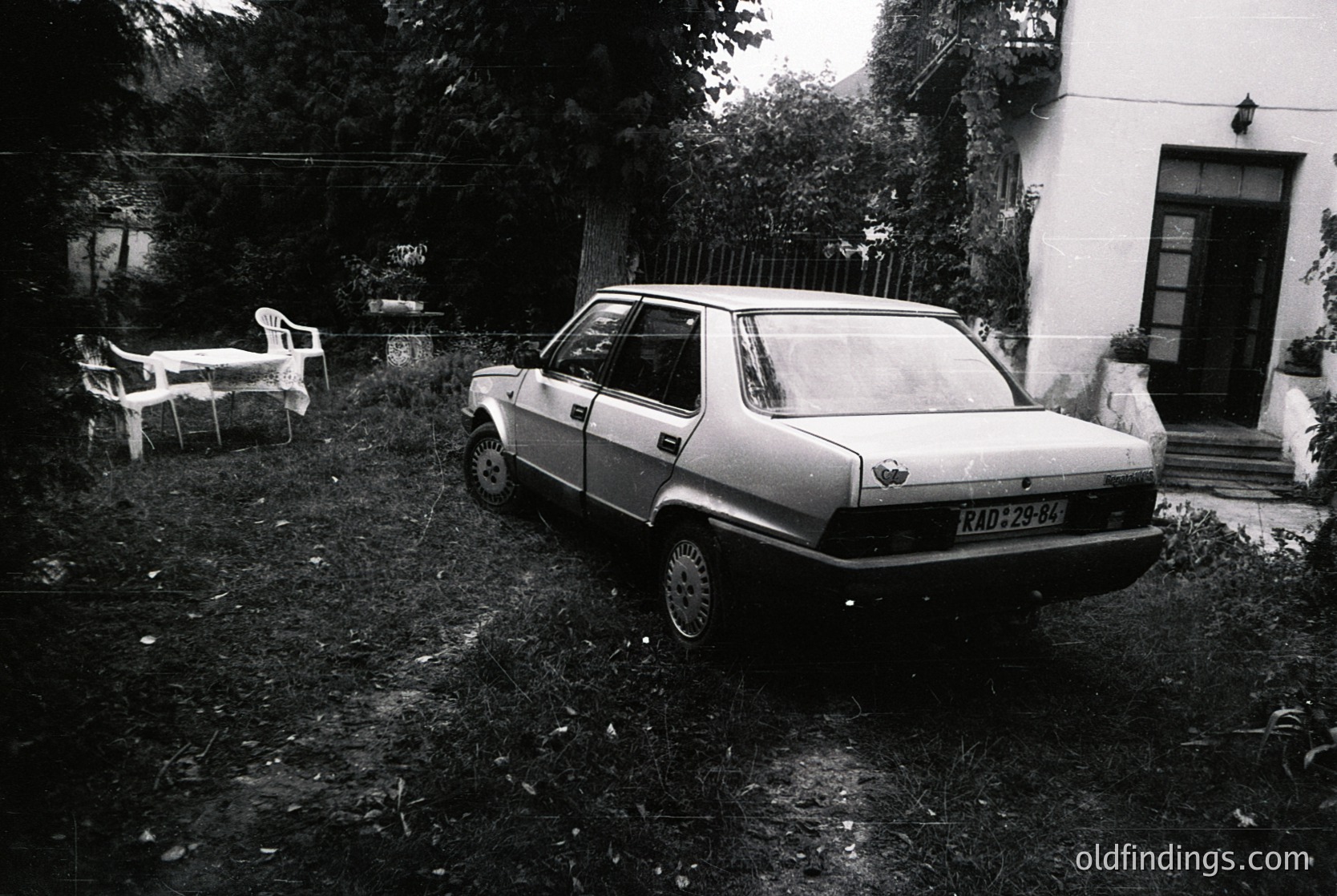 Vintage sedan parked in overgrown backyard with faded white plastic chair and table. Rustic residential setting, likely Eastern Bloc-era due to car design.