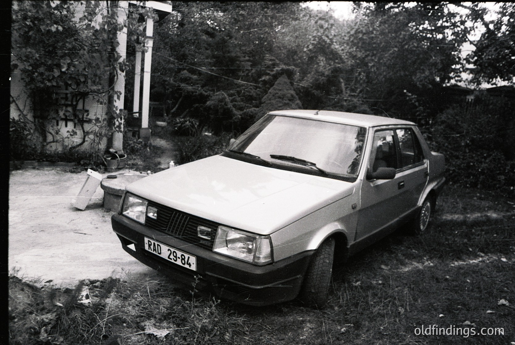 Vintage sedan (likely Škoda 130/136) parked on overgrown concrete, license plate "ВАД 29-84" suggests Bulgarian registration. Rusting concrete wall and weathered trees indicate mid-20th century urban decay.