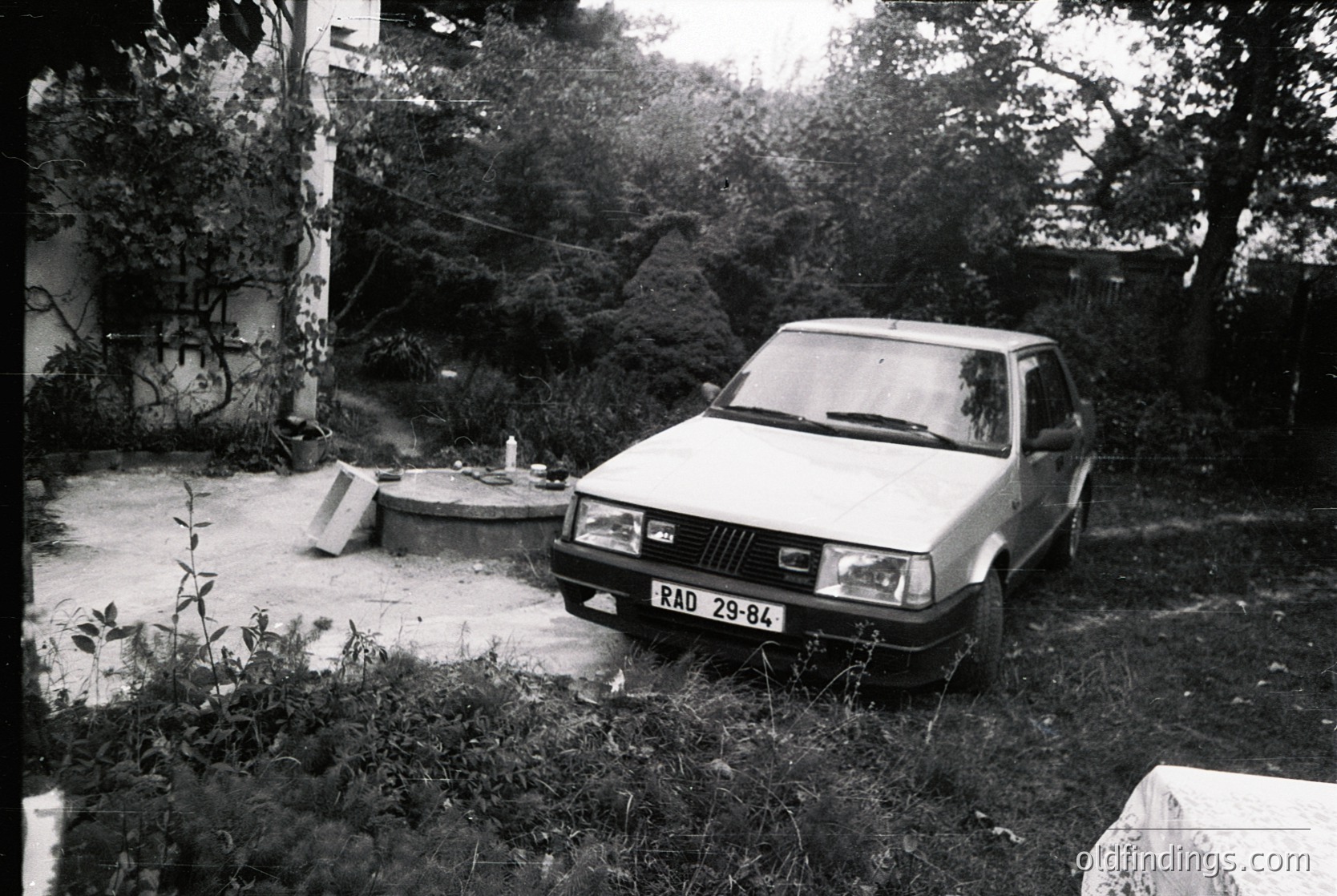 Vintage sedan parked on overgrown concrete slab beside concrete well, surrounded by dense foliage and graffiti on adjacent wall. Likely Eastern Bloc-era vehicle ( 29-84 license plate). Urban decay aesthetic with mid-century architecture.