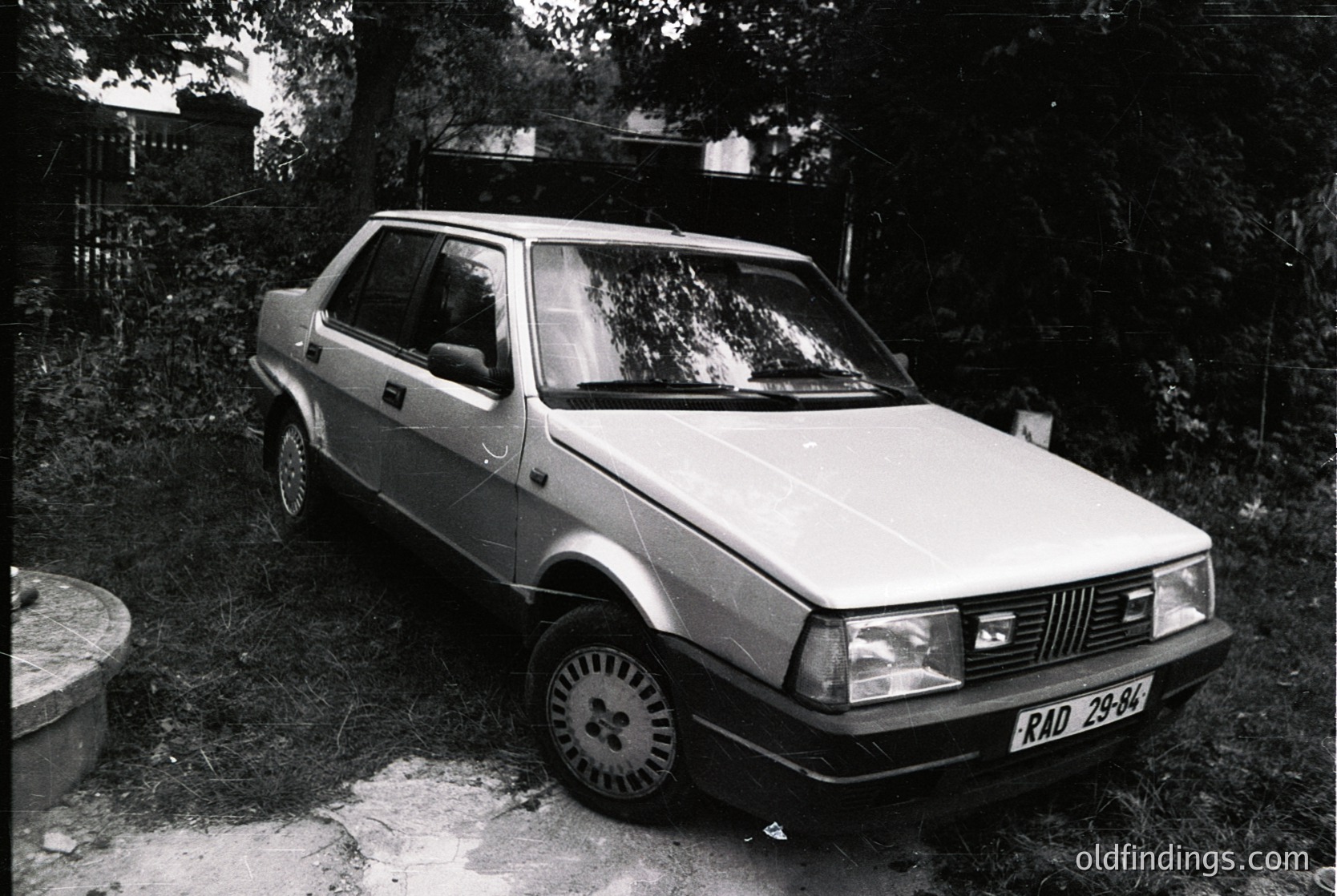 Classic hatchback sedan parked in a wooded area, likely a Fiat 127 based on grille design. Black-and-white photo captures 1970s–1980s era with visible license plate "RAD 29-84." Rust and weathering suggest vintage condition. Ideal for automotive history or stock photography.