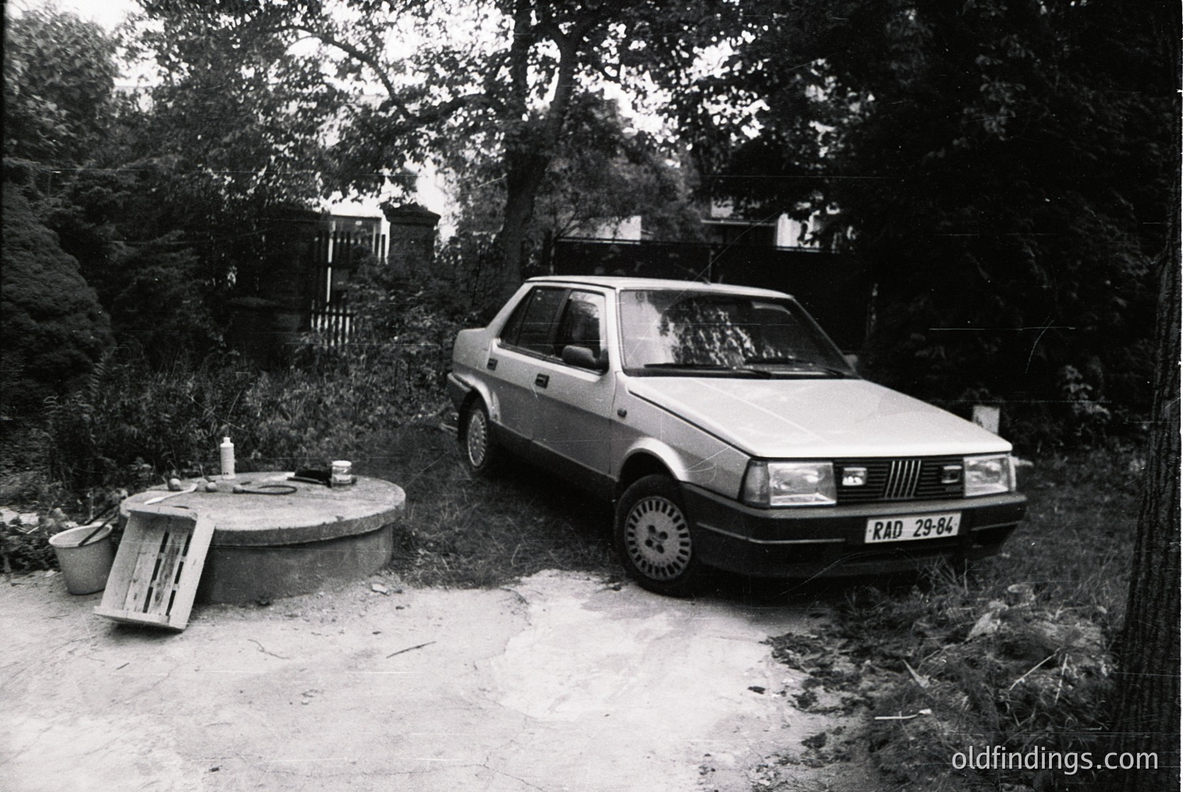 1970s-era Fiat 128 parked beside a rustic concrete picnic table and metal bucket. Dense forest and rocky terrain suggest a rural or mountainous setting. Vintage license plate (RAD 29-84) indicates Eastern Bloc region. Ideal for automotive history, vintage travel, or nostalgic design references.