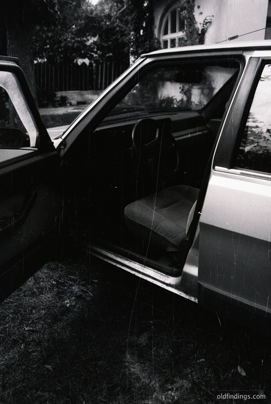 Classic mid-century station wagon interior, likely a **Volkswagen Type 4 (Bus)** or similar 1960s-70s model. Open door reveals worn vinyl seats, exposed metal frame, and rearview mirror. Rain-soaked pavement and blurred greenery suggest a residential or suburban setting. Ideal for vintage automotive, transportation history, or nostalgic design references.
