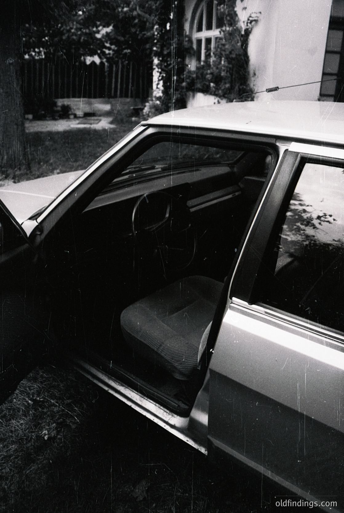 Classic sedan interior captured mid-rain, interior light reflecting off wet surfaces. Vinyl bucket seat, round instrument cluster, and chrome door handle visible. Residential driveway with brick path and greenery in background. Likely late 20th-century European design.