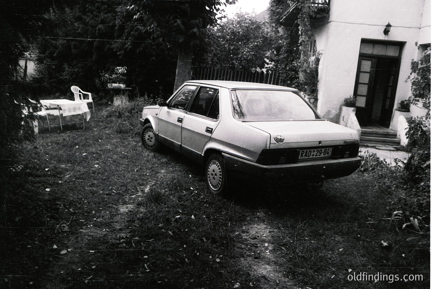 Vintage sedan parked on overgrown grass beside a residential building, likely Eastern European . Distinctive rear license plate "RAD-29-84" suggests Bulgarian registration. White plastic chair and faded sign in background hint at rural or suburban setting. Ideal for automotive history, vintage travel, or cultural documentation.