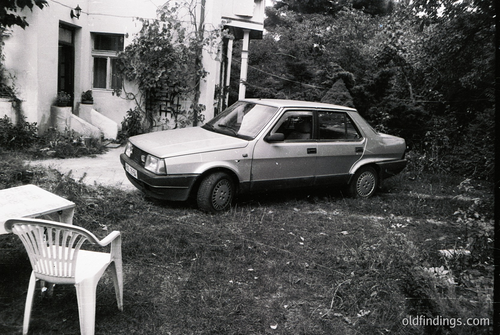 Vintage sedan parked on overgrown grass beside a white plastic chair and 1970s-era building with arched windows. Rustic, mid-century European residential setting.