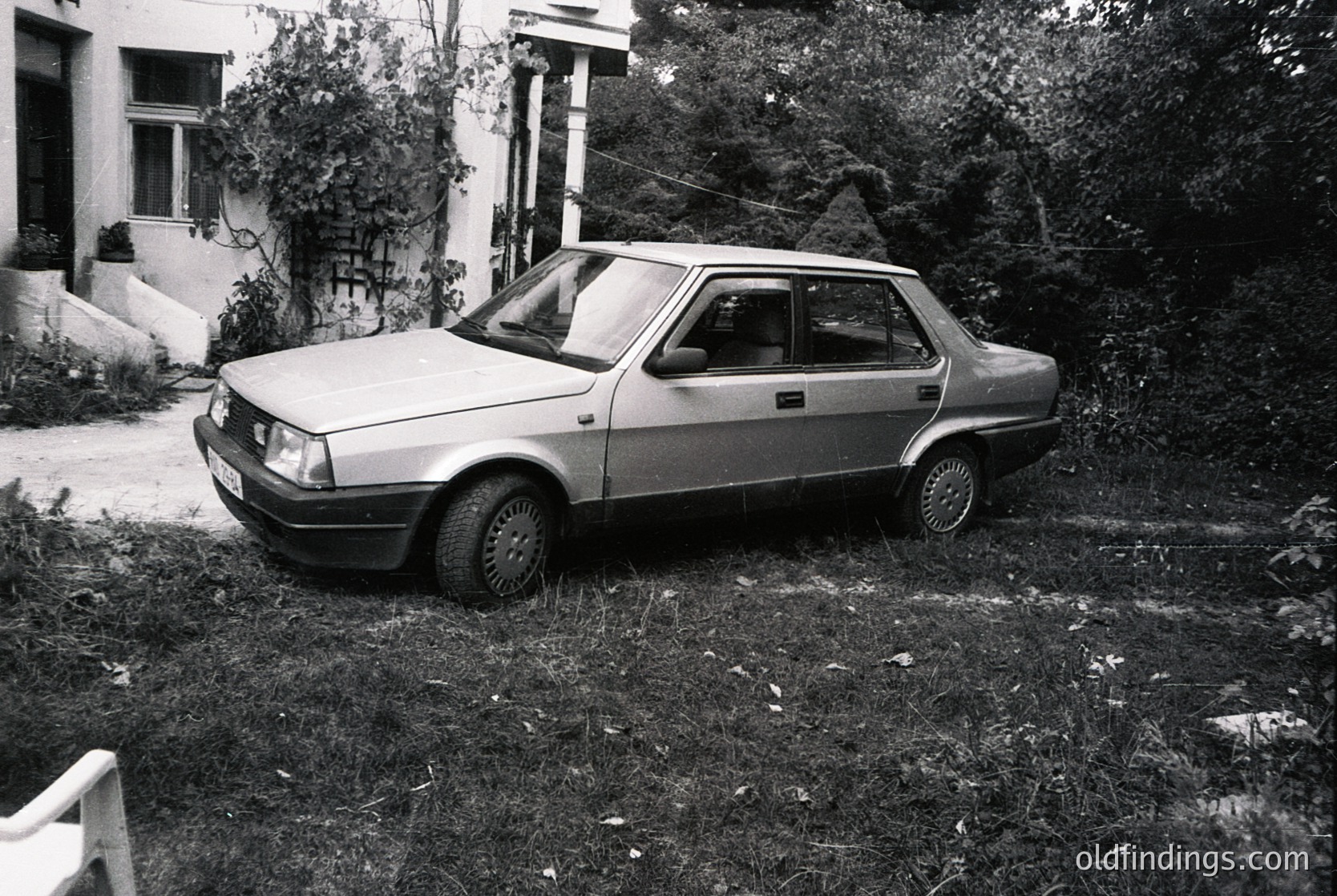 Vintage sedan parked on overgrown grass beside a residential driveway, likely a 1970s–1980s European hatchback. Noteworthy details: hubcaps, minimalist rear spoiler, and a partially visible rear license plate. Surrounding architecture includes a white-painted house with columns and a gated entrance.