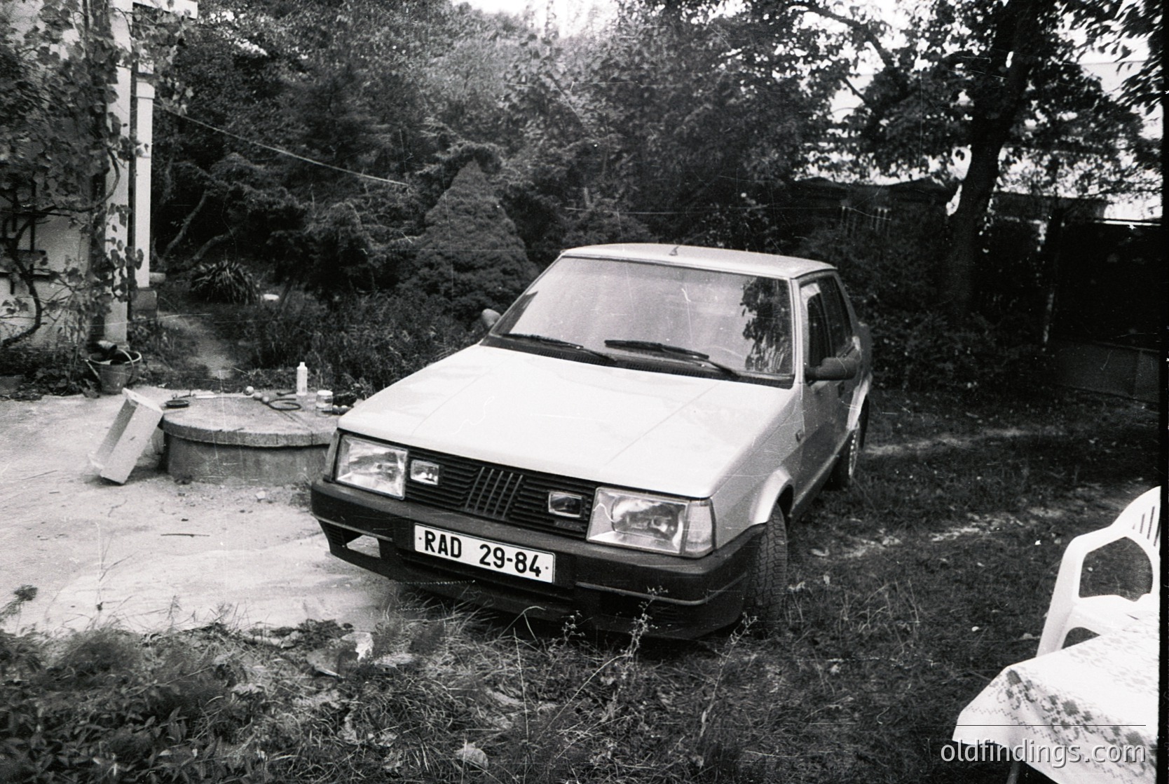 1980s-era Fiat sedan (RAD 29-84) parked in overgrown backyard, surrounded by concrete slab and rusted metal barrel. Residential setting with dense foliage and weathered concrete structures. Black-and-white photo captures mid-20th-century European domestic life.
