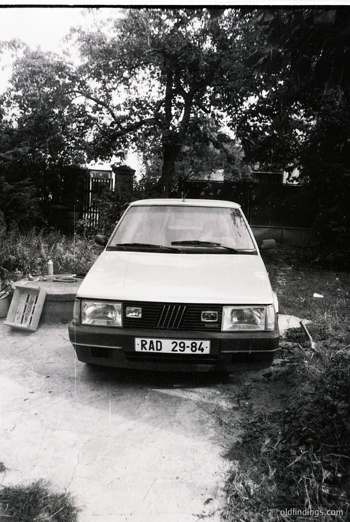 1980s-era hatchback sedan (likely a Fiat 127 or similar) parked on a gravel driveway, identifiable by the "RAD 29-84" license plate. The car’s design suggests Eastern European production. Surrounding foliage and weathered wooden structures hint at a rural or suburban setting.