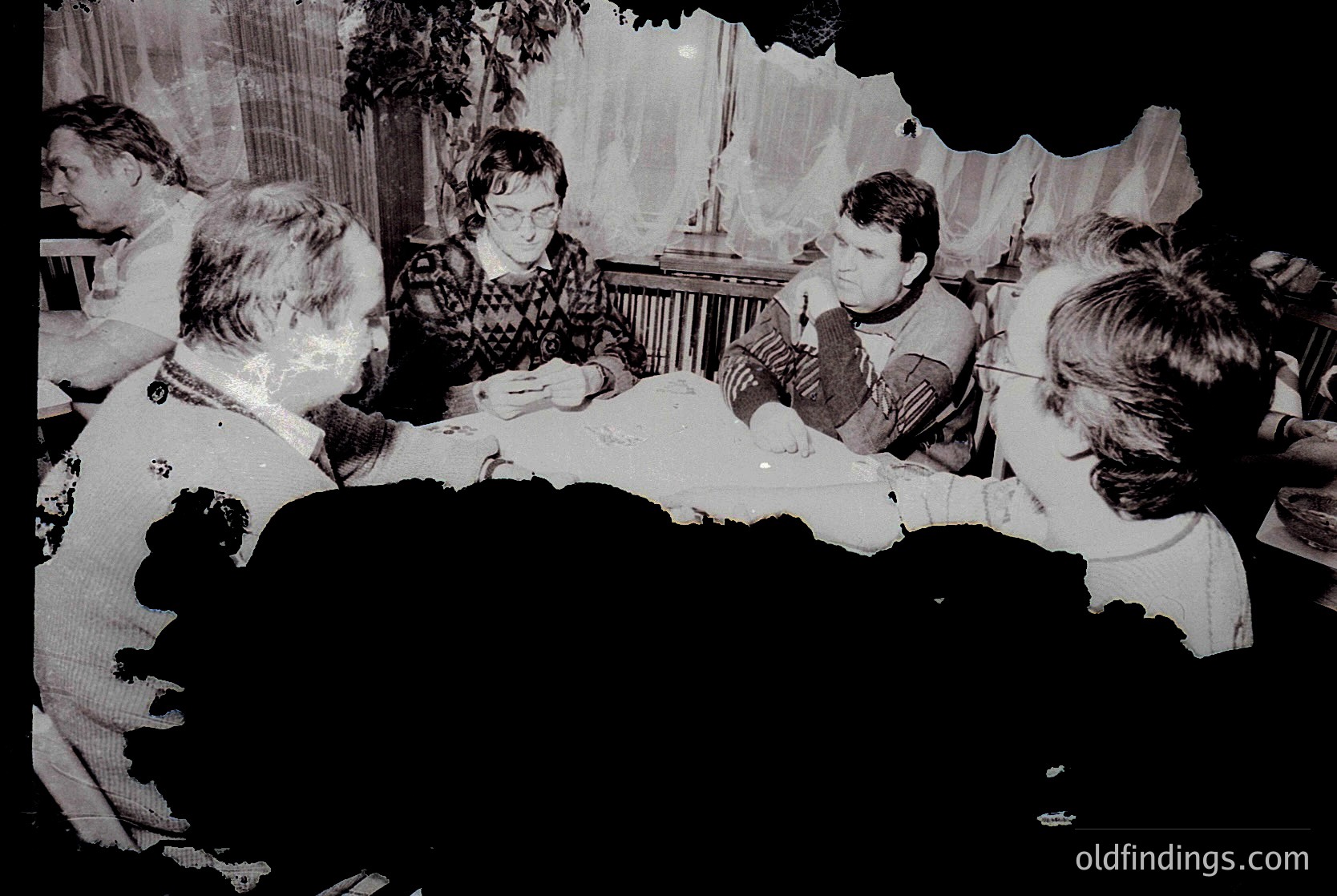 Vintage black-and-white photo of a group seated around a round table in a dimly lit indoor setting, likely a 1960s-70s gathering. Visible details include patterned tablecloth, a central object resembling a cake or decorative item, and attendees in casual, mid-century attire. The blurred background suggests a cozy, possibly residential or community hall atmosphere.