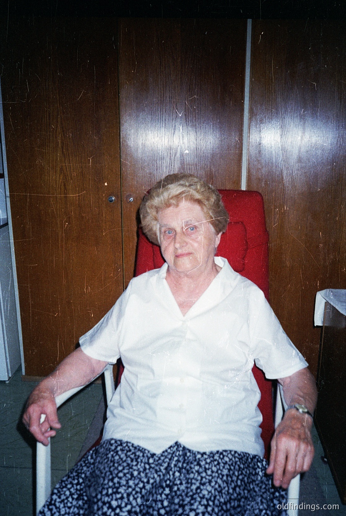 Portrait of an elderly woman seated in a red upholstered armchair, holding armrests for support. She wears a white blouse and patterned pants, with a wristwatch visible. The setting appears to be an institutional or residential interior, likely from the mid-20th century. [mid20thcenturyportrait ]
