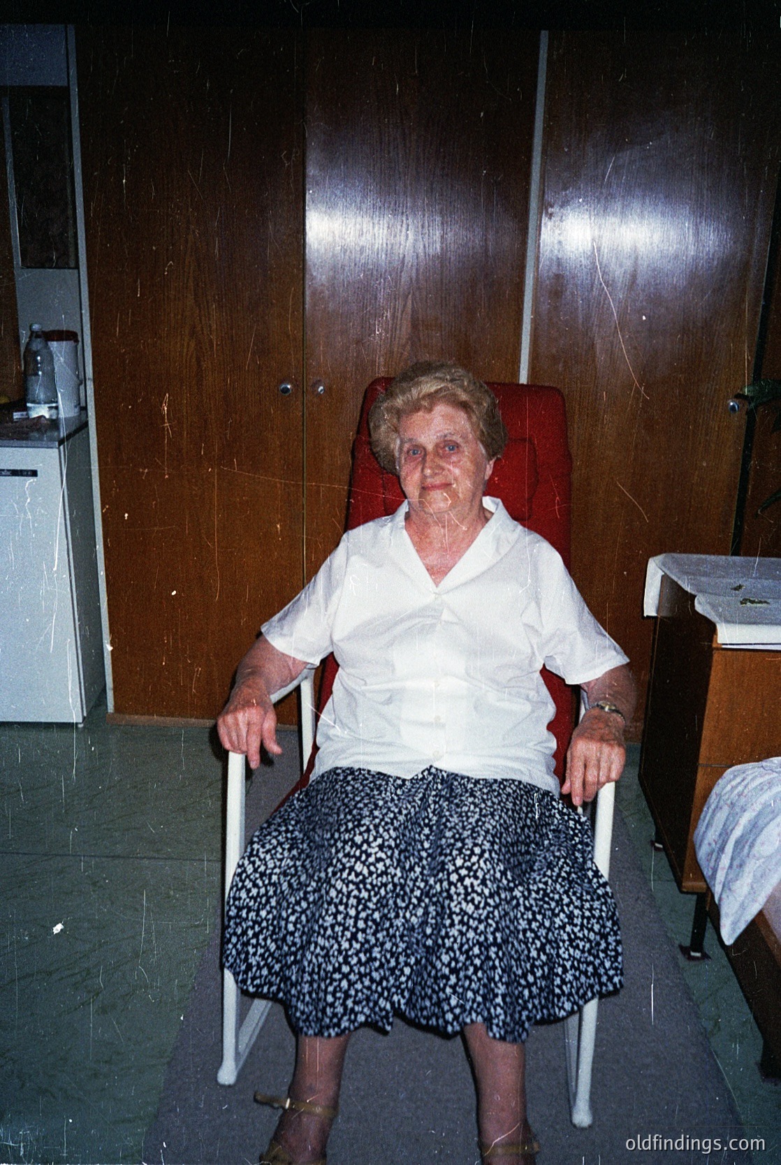 Mid-20th century indoor portrait of an elderly woman in a wheelchair, wearing a white blouse and patterned skirt. Wood-paneled walls and a vintage cabinet with a stove in the background suggest a residential or institutional setting, likely Eastern Europe. The lighting and styling indicate a posed studio or home portrait from the 1960s–1970s.