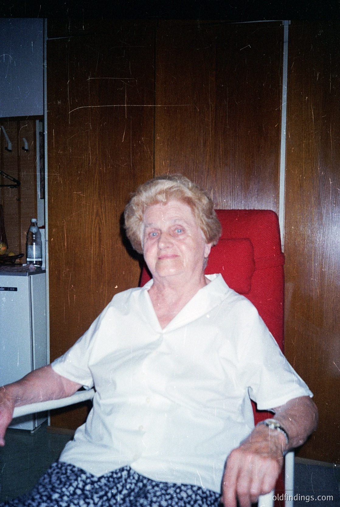 Portrait of an elderly woman seated in a modest indoor setting, likely mid-20th century. She wears a light-colored blouse and patterned pants, with a watch on her wrist. Behind her, a red chair and wooden cabinet suggest a lived-in home. The image evokes mid-century domestic life.
