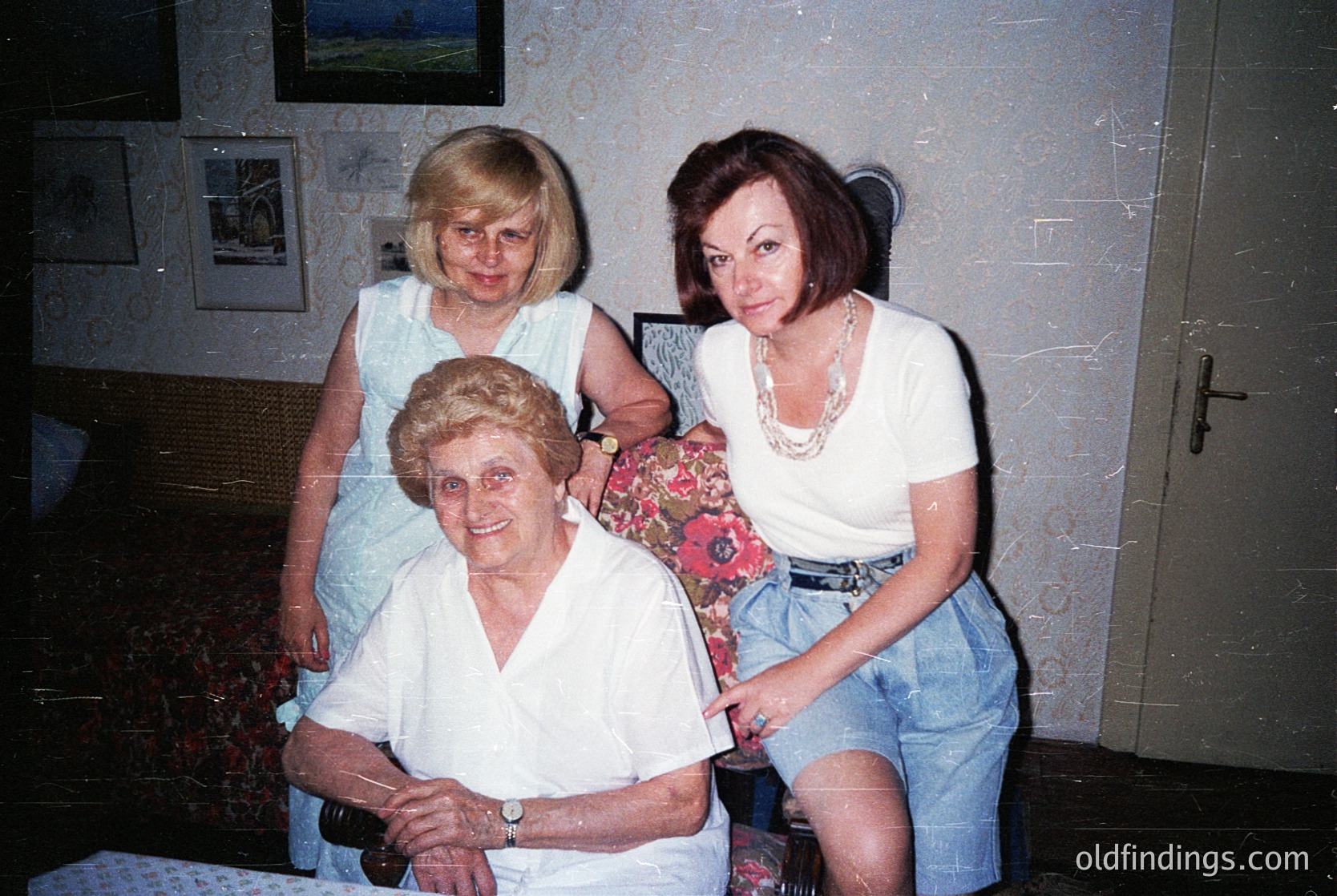 Three generations pose indoors, likely 1980s–1990s. Elderly woman seated in vintage armchair, flanked by a middle-aged woman in floral blouse and a younger woman in denim shorts and layered necklaces. Wall-mounted framed photos and patterned wallpaper suggest a lived-in, familial setting.