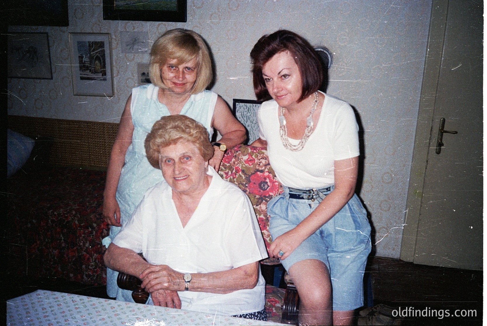 Three generations of women pose indoors, likely Eastern Europe, late 20th century. The seated woman wears a vintage white blouse with a watch, flanked by a middle-aged woman in a floral top and a younger woman in denim shorts and a layered necklace. Framed photos on the wall suggest a personal, domestic setting. Warm lighting and patterned wallpaper hint at a mid-century interior.