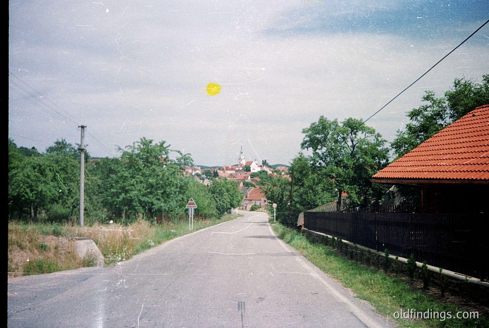 Vintage rural road scene with lush greenery, narrow asphalt path, and distant village rooftops. Sun flare highlights midday lighting. Black fence and utility pole on right. Likely Eastern European countryside, 1980s–1990s.