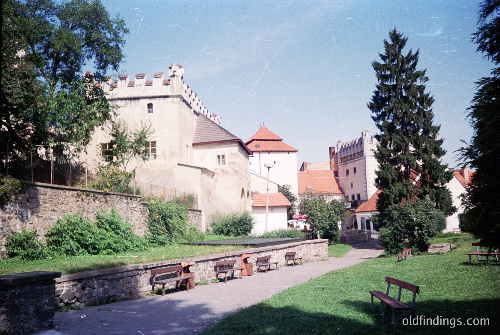 Medieval castle complex with stone towers, turrets, and fortified walls, surrounded by lush greenery. Prominent wooden benches along a paved pathway. Likely Eastern European architecture, possibly or . Mid-20th century (1950s–1970s) based on vegetation and structure condition.