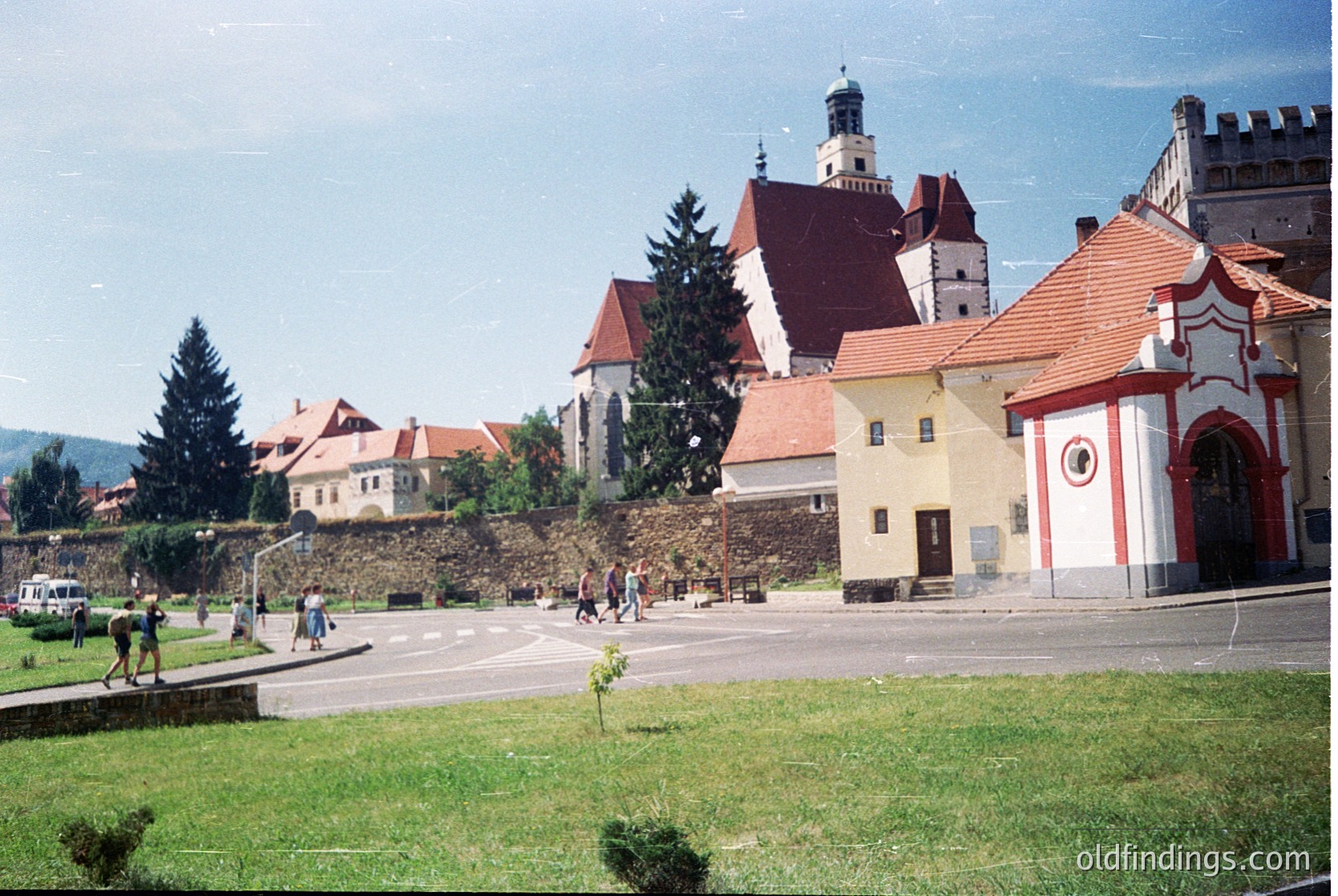 Historic European town square with medieval architecture—red-tiled roofs, stone walls, and a prominent church with a bell tower. Lush greenery and a few pedestrians suggest a preserved heritage site. Likely late 20th century.