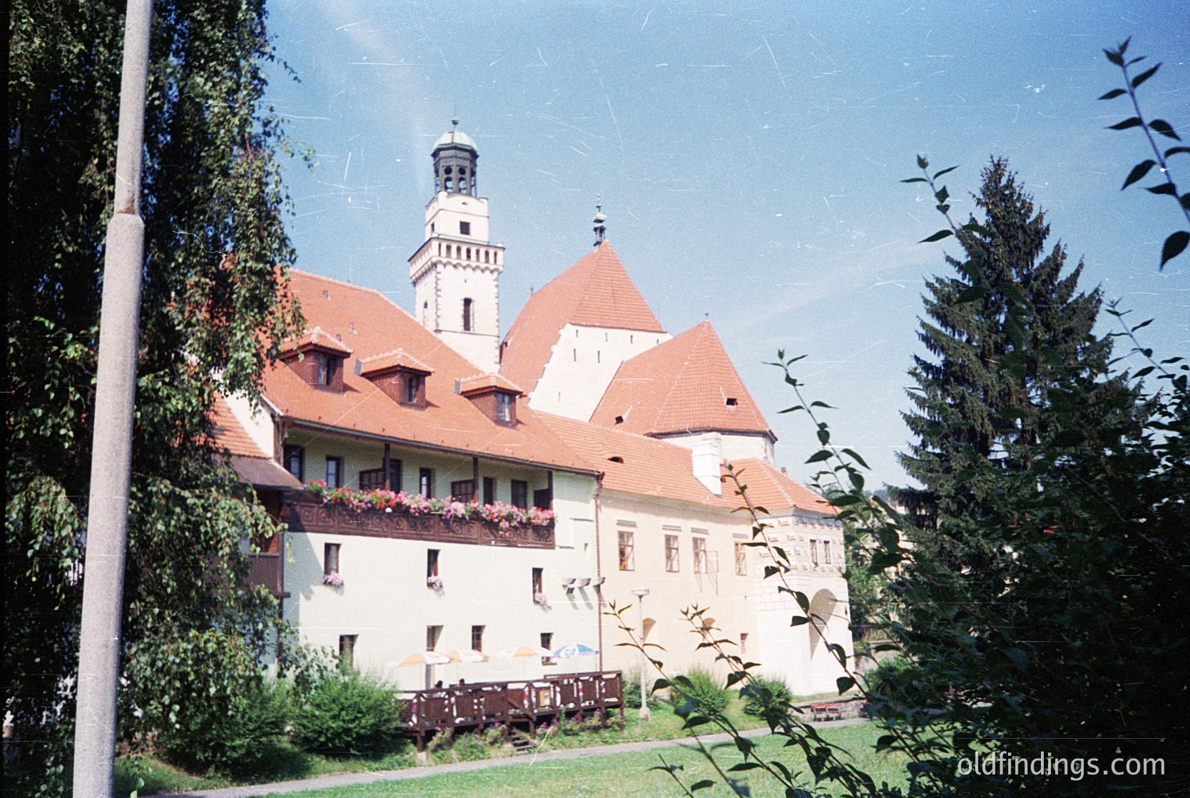 Historic castle with red-tiled gables and central tower, surrounded by lush greenery. Likely European architectural style, possibly 18th–19th century. Symmetrical façade with flower boxes under windows.