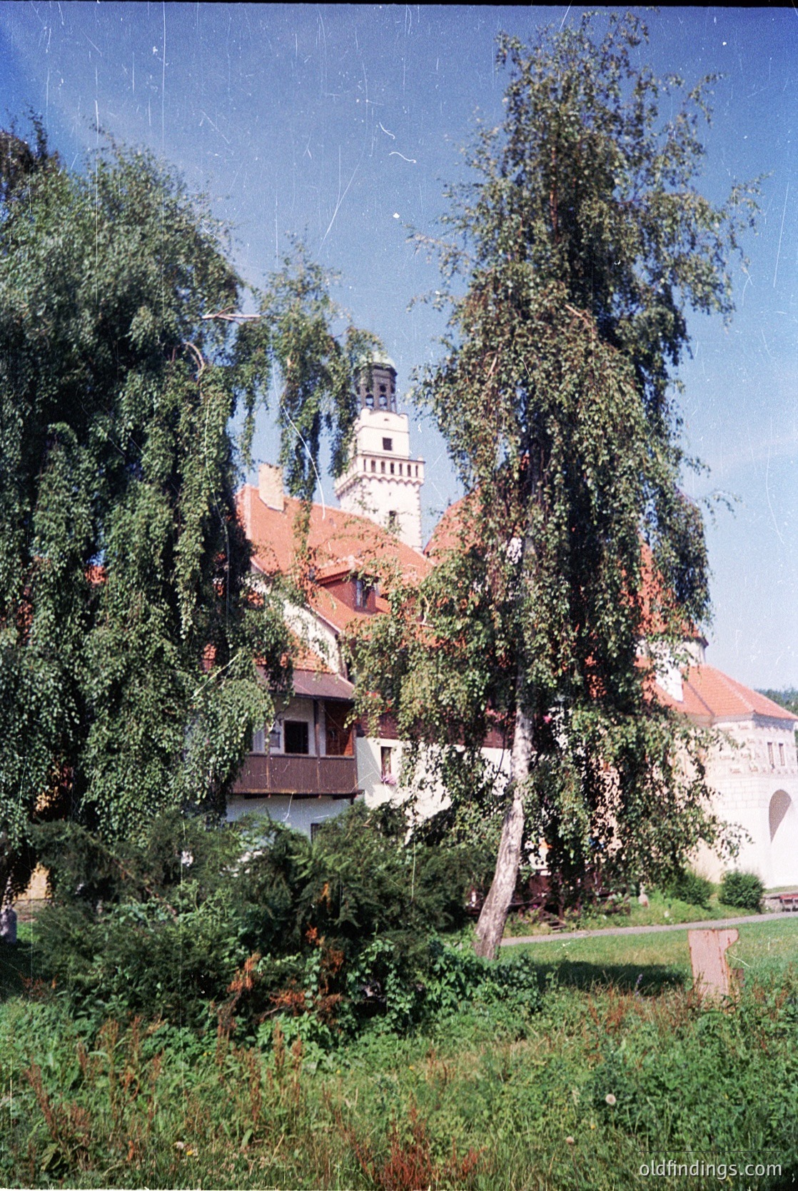 Historic European villa with red-tiled roof and white walls, framed by dense greenery. Central tower with arched windows suggests 19th-century architectural style. Lush garden and mature trees enhance the serene, rural setting.