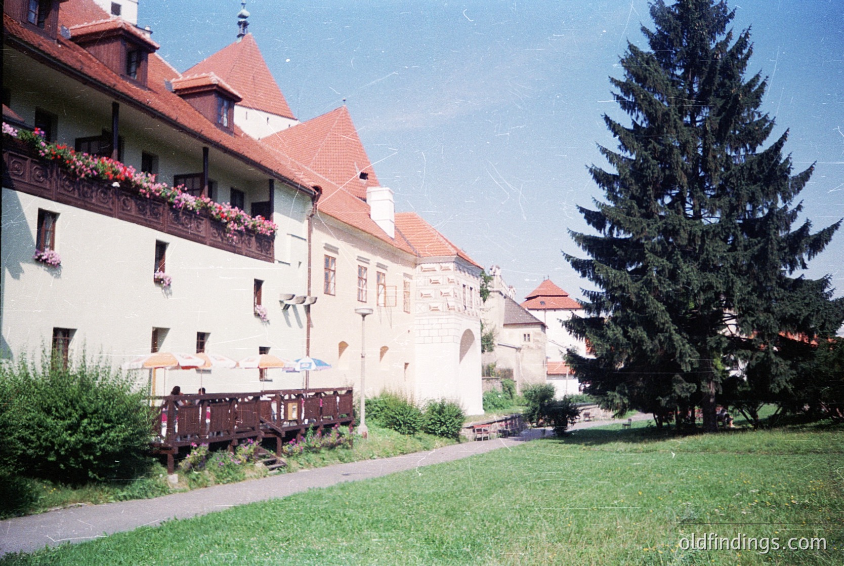 Vintage European courtyard with cream-colored building featuring red-tiled gables, wrought-iron balconies, and blooming flowers. Wooden picnic tables and benches line a paved pathway under a tall evergreen. Likely 1960s–1980s era, suggesting a preserved historic town center.