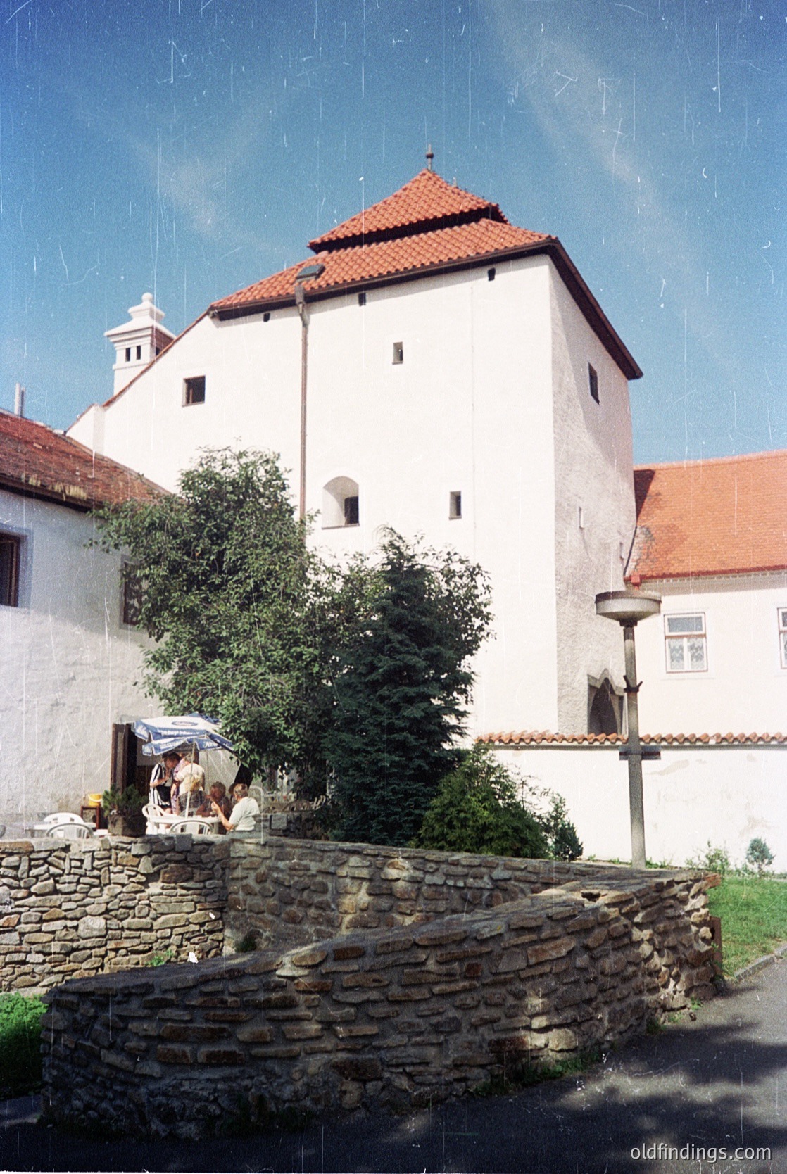 Historic stone-walled courtyard with a prominent medieval-style tower featuring red-tiled conical roof and small arched windows. Adjacent whitewashed building with red-tiled gables and a chimney. Stone retaining wall with greenery and a small outdoor seating area under a canopy. Likely Eastern European architectural style.