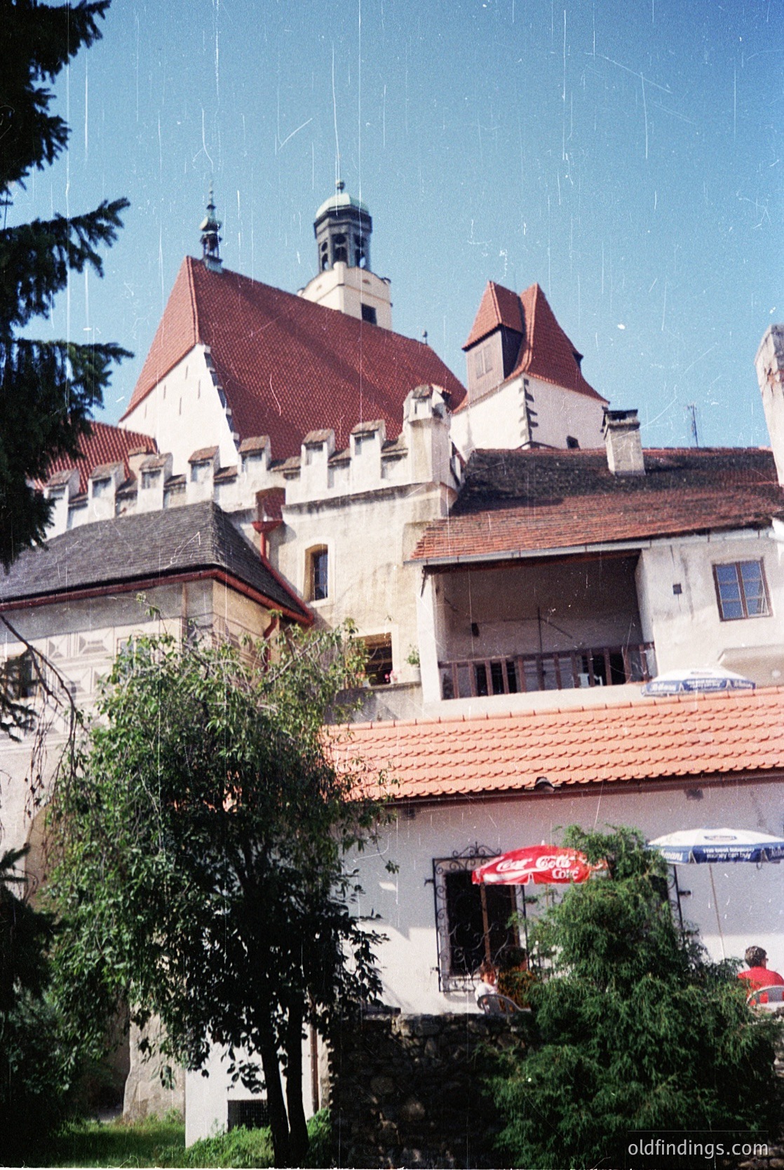 Historic castle complex with Gothic-Renaissance architecture—steepled towers, crenellated parapets, and red-tiled roofs. Outdoor seating with red umbrellas suggests modern café use. Lush greenery frames the scene, hinting at a European setting. Likely –16th century, possibly () or similar medieval region.