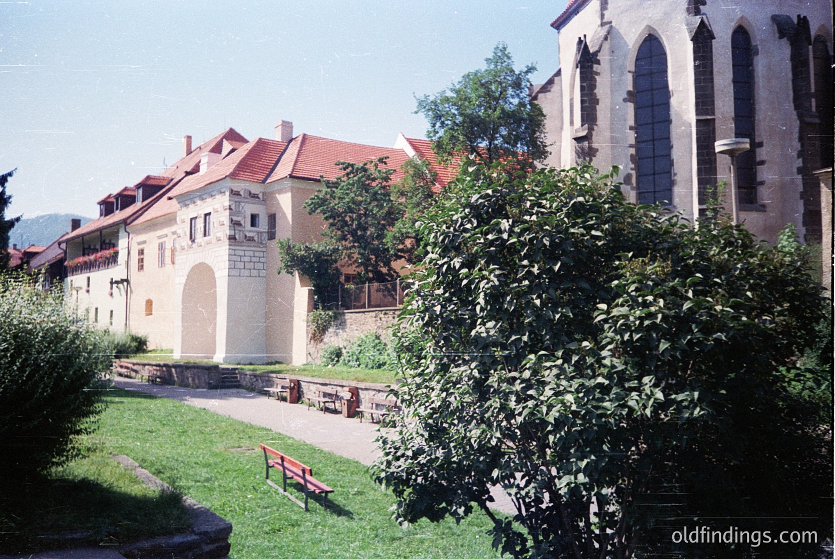 Historic courtyard featuring a stone archway entrance flanked by red-tiled buildings. Gothic-style church with pointed arches and arched windows in background. Lush greenery and benches suggest a serene public space. Likely Eastern European architectural style.