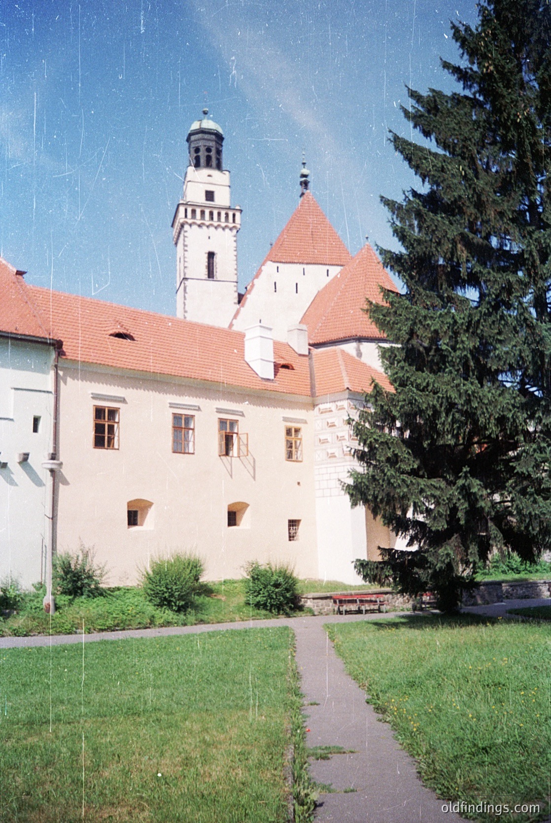 Historic castle complex with red-tiled gables and a central tower, likely European . Symmetrical façade with small rectangular windows and a paved courtyard path. Lush greenery and evergreen trees frame the scene, suggesting a preserved heritage site. Potential or earlier origin.