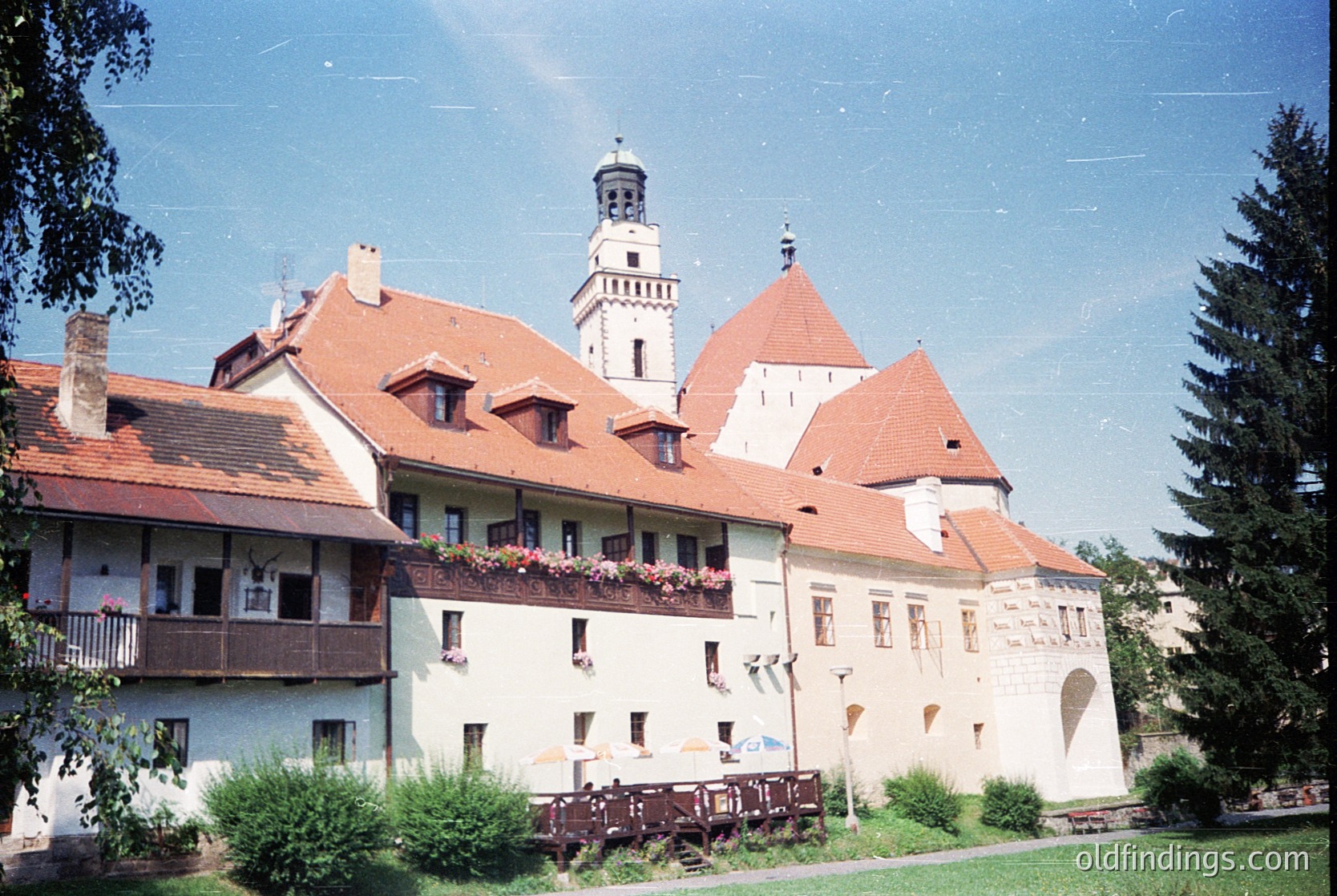 Historic European castle complex with red-tiled roofs and white plaster walls, featuring a central tower with crenellations. Balconies with flower boxes and laundry hanging suggest residential use. Lush greenery and mature trees frame the scene, indicating a well-preserved heritage site. #