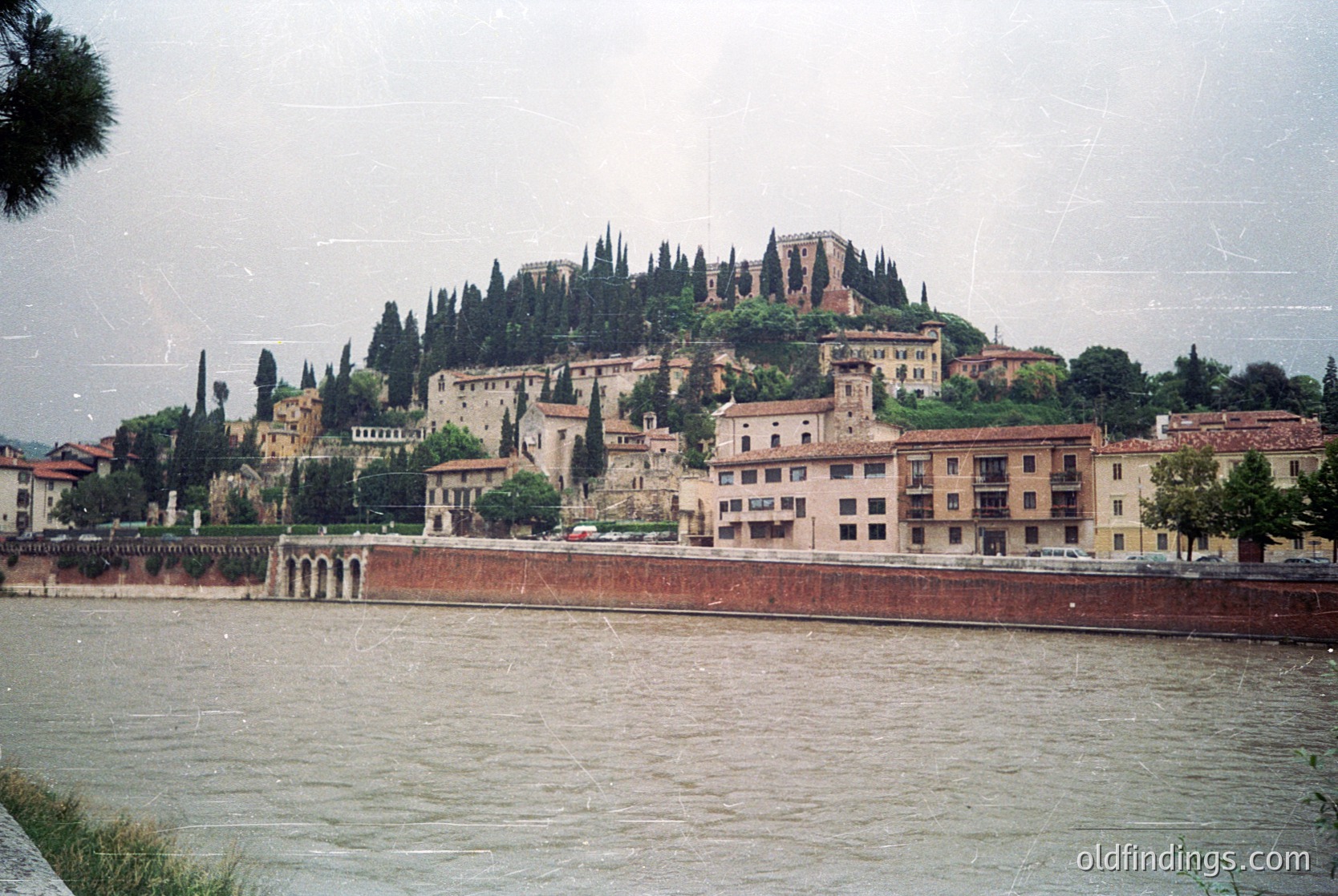 Historic hilltop fortress perched atop cypress-lined slopes, overlooking a riverbank lined with mid-20th-century buildings. Stone walls and arched bridges frame the scene, suggesting a fortified medieval origin. Likely , Italy, with -era urban development in foreground.