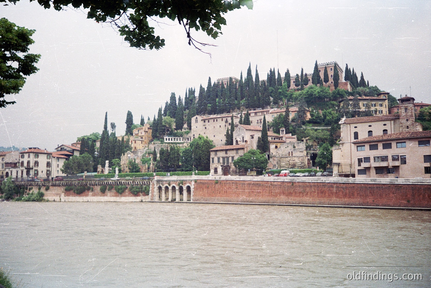 Historic riverside town with medieval hilltop fortress and cypress-lined slopes. Stone buildings and arched bridges frame the Adige River, suggesting Verona, Italy. Overcast lighting enhances atmospheric depth.