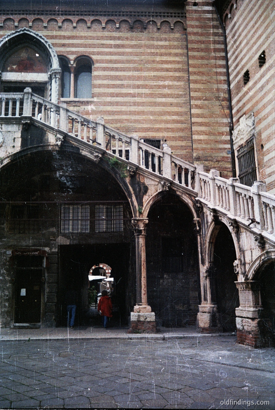 Renaissance-era stone courtyard with arched walkways and spiral staircase in Venice, Italy. Weathered brick walls and intricate stonework highlight historic Venetian architecture. Single figure in red coat walking beneath arched entrance ( )