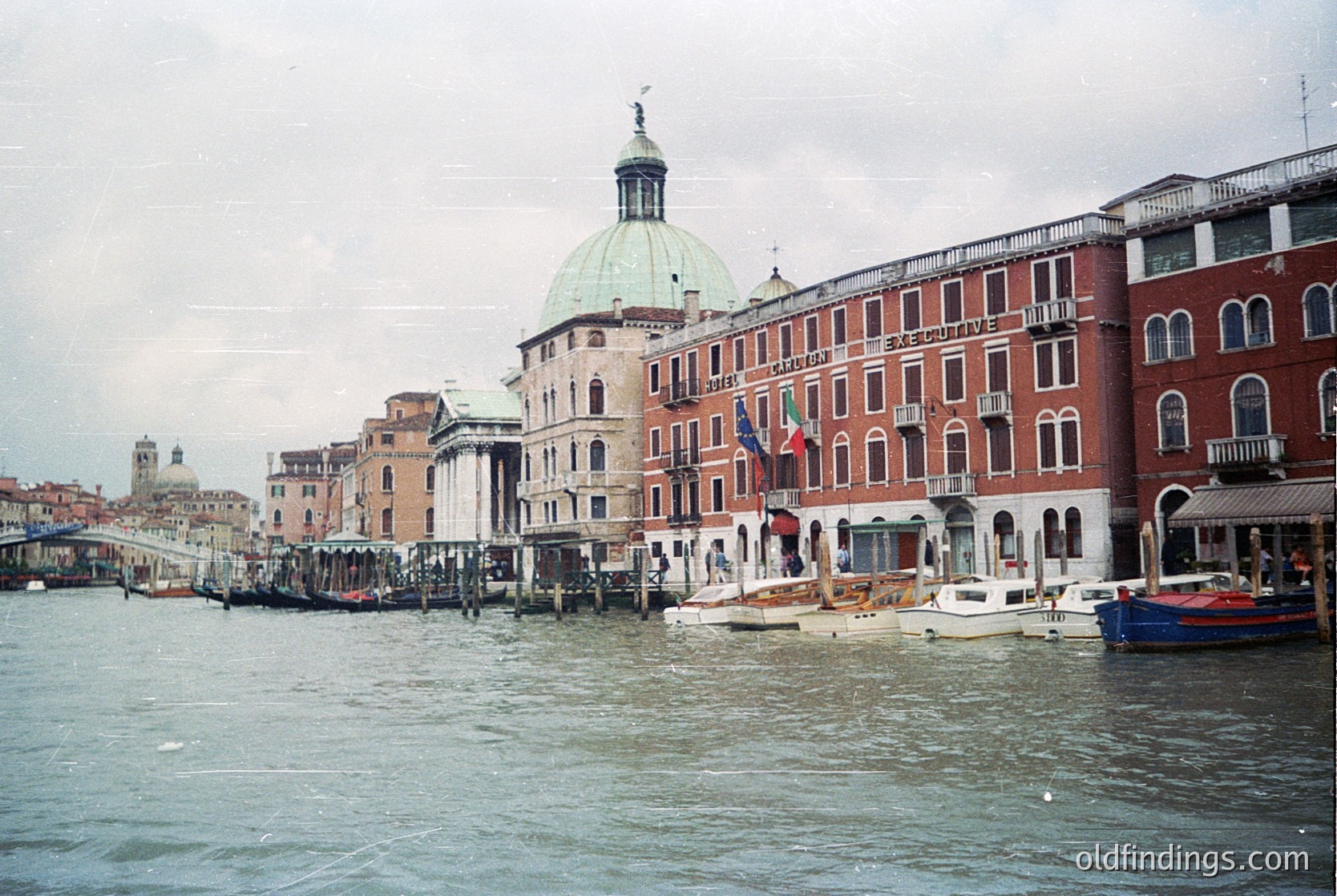 Venetian canal scene featuring historic architecture: domed church with classical façade, adjacent brick-lined buildings, and moored gondolas. Overcast sky enhances muted tones. Likely late 20th century.